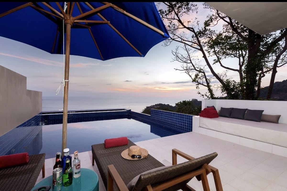 An infinity pool with a view of the sunset, a white sofa with red cushions, and a table with drinks and a hat.