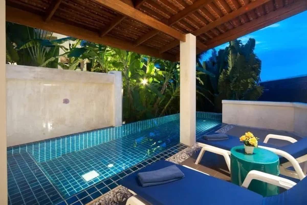 A covered outdoor pool area with blue-tiled flooring, lounge chairs, and a small table with a flower vase.