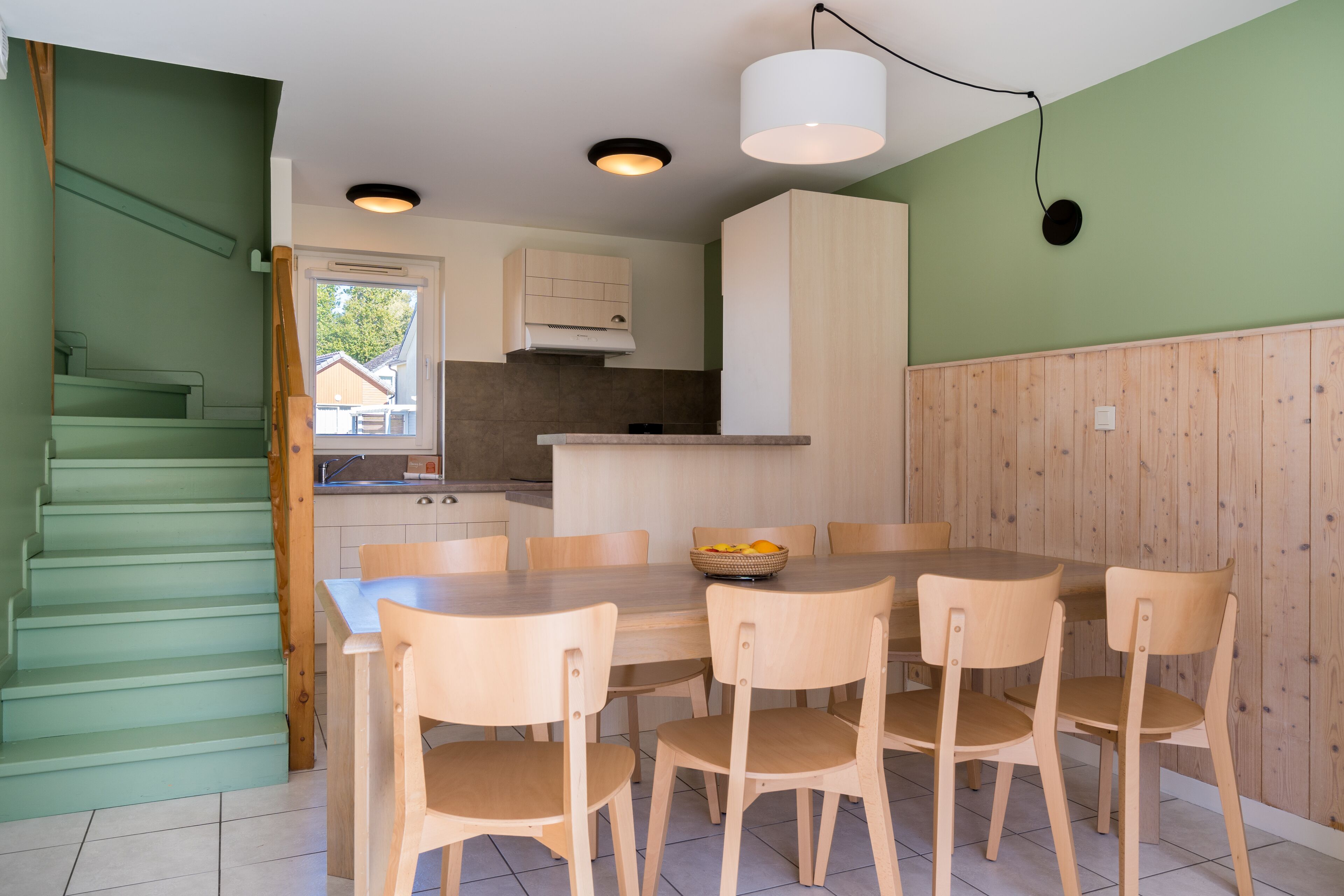 A modern kitchen with wooden cabinets, a dining table with chairs, and a staircase with green walls.