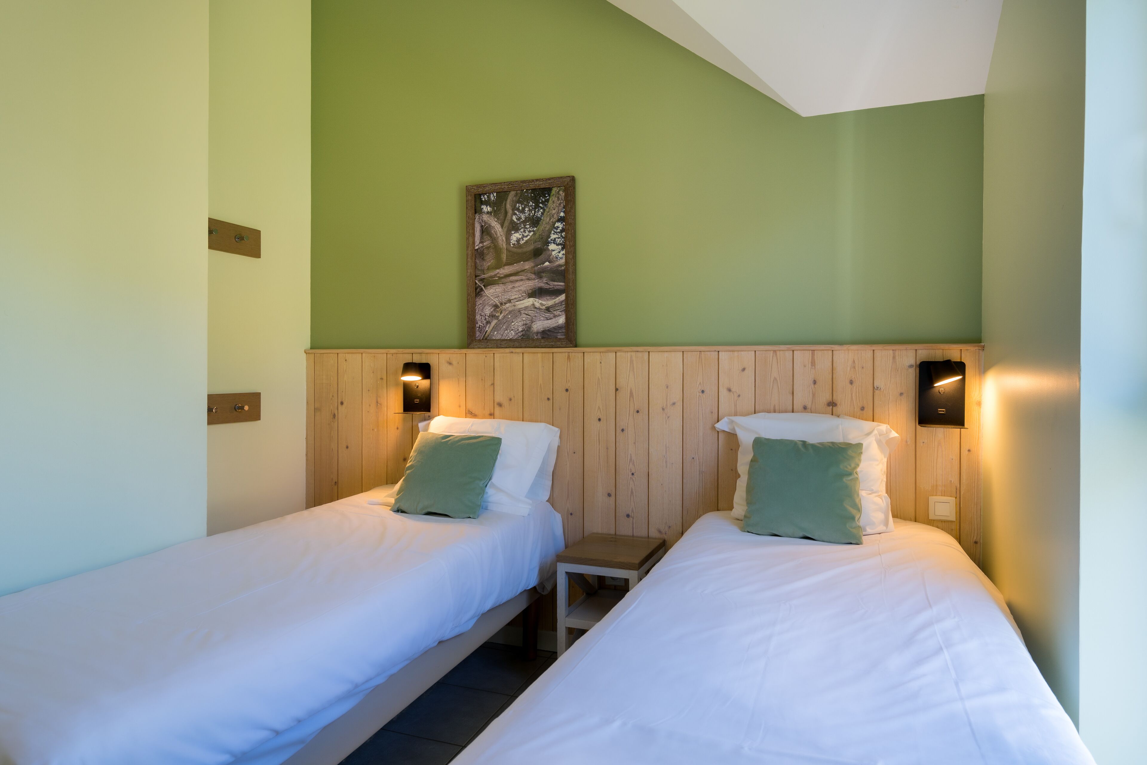 Ein Zimmer mit zwei Einzelbetten, jeweils mit Kissen und einem kleinen Nachttisch. Über jedem Bett befindet sich eine Wandleuchte.