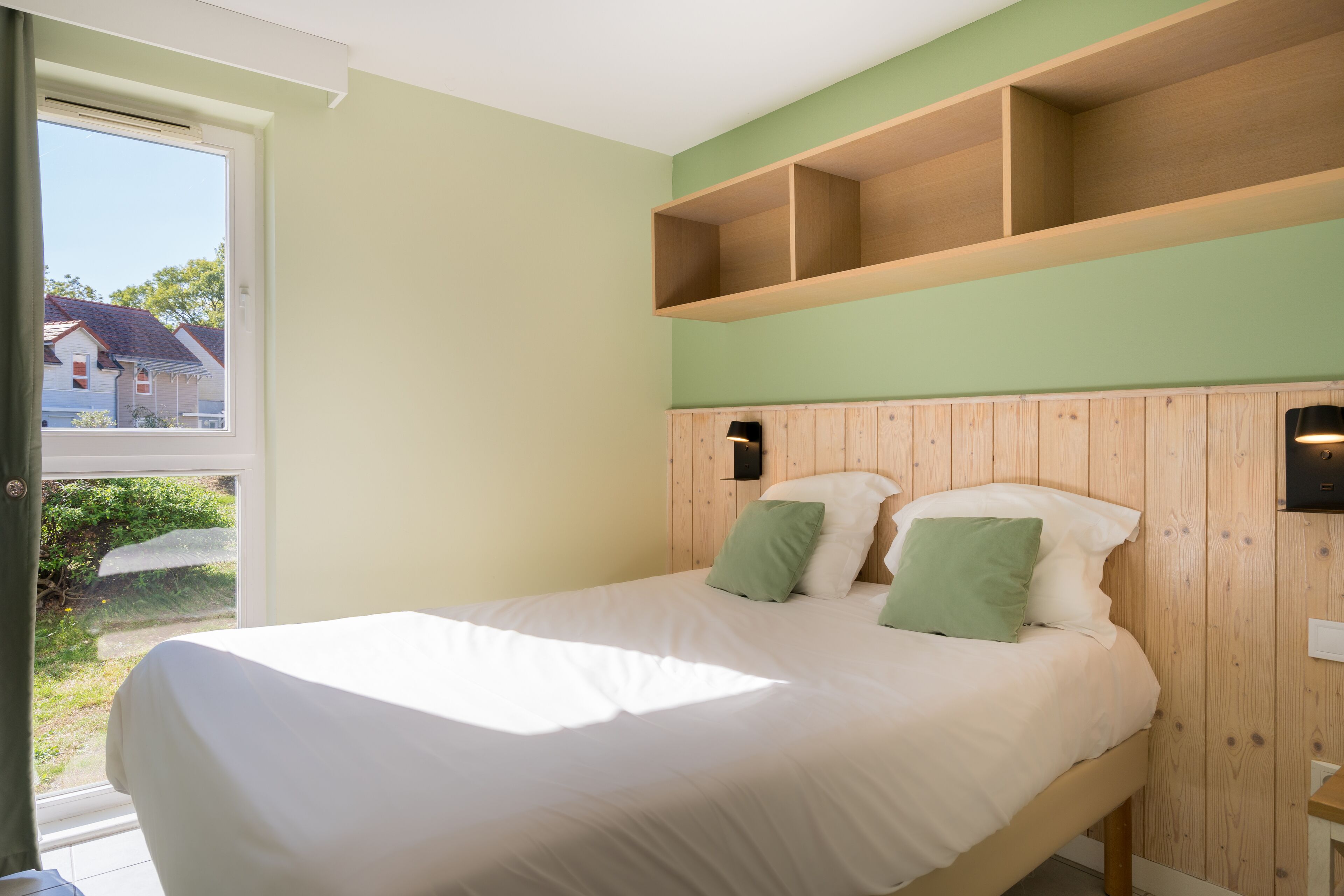 A bedroom with a bed, wooden headboard, and a window with a view of houses.