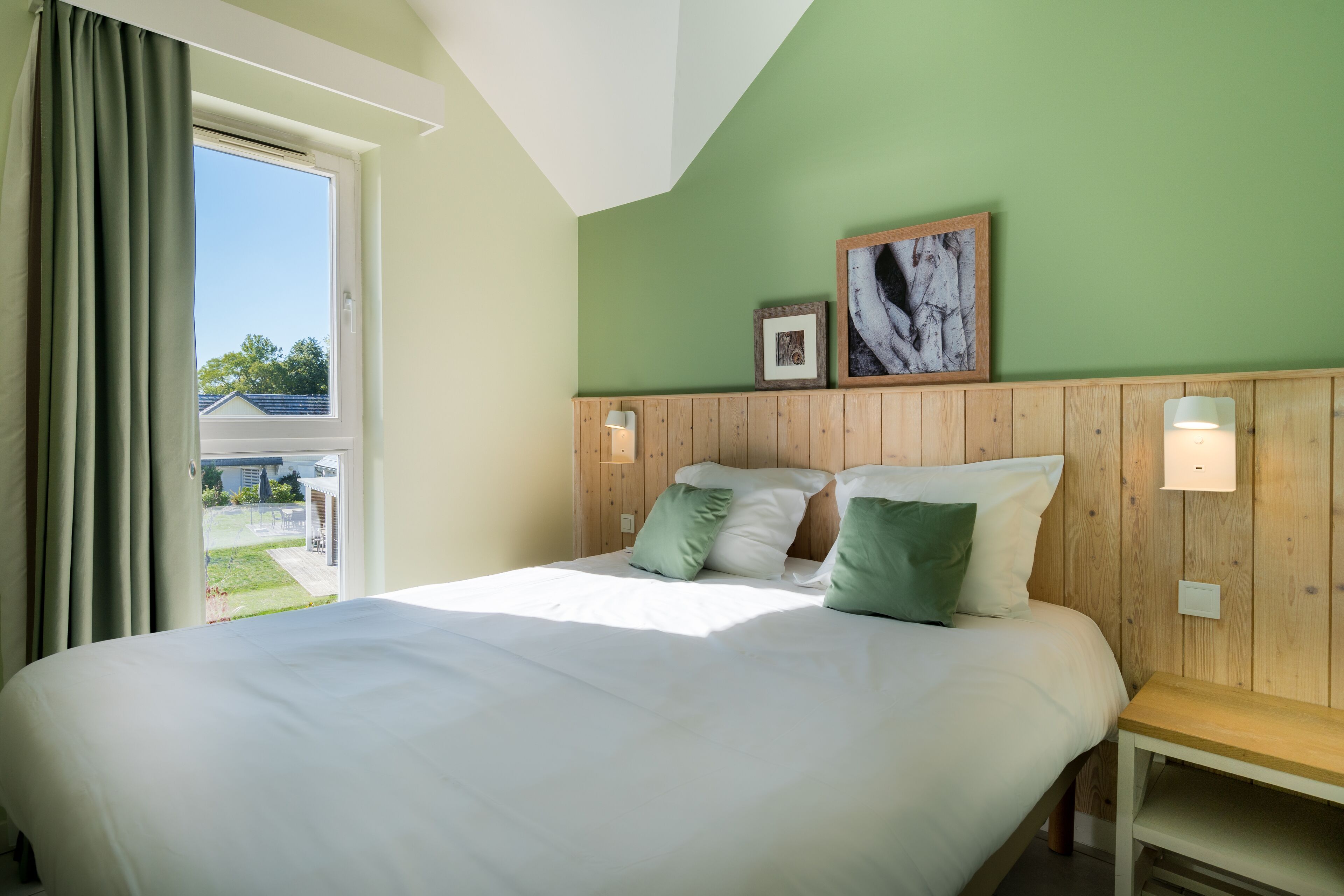 Quarto com cama grande, cabeceira de madeira e vista para um jardim pela janela.