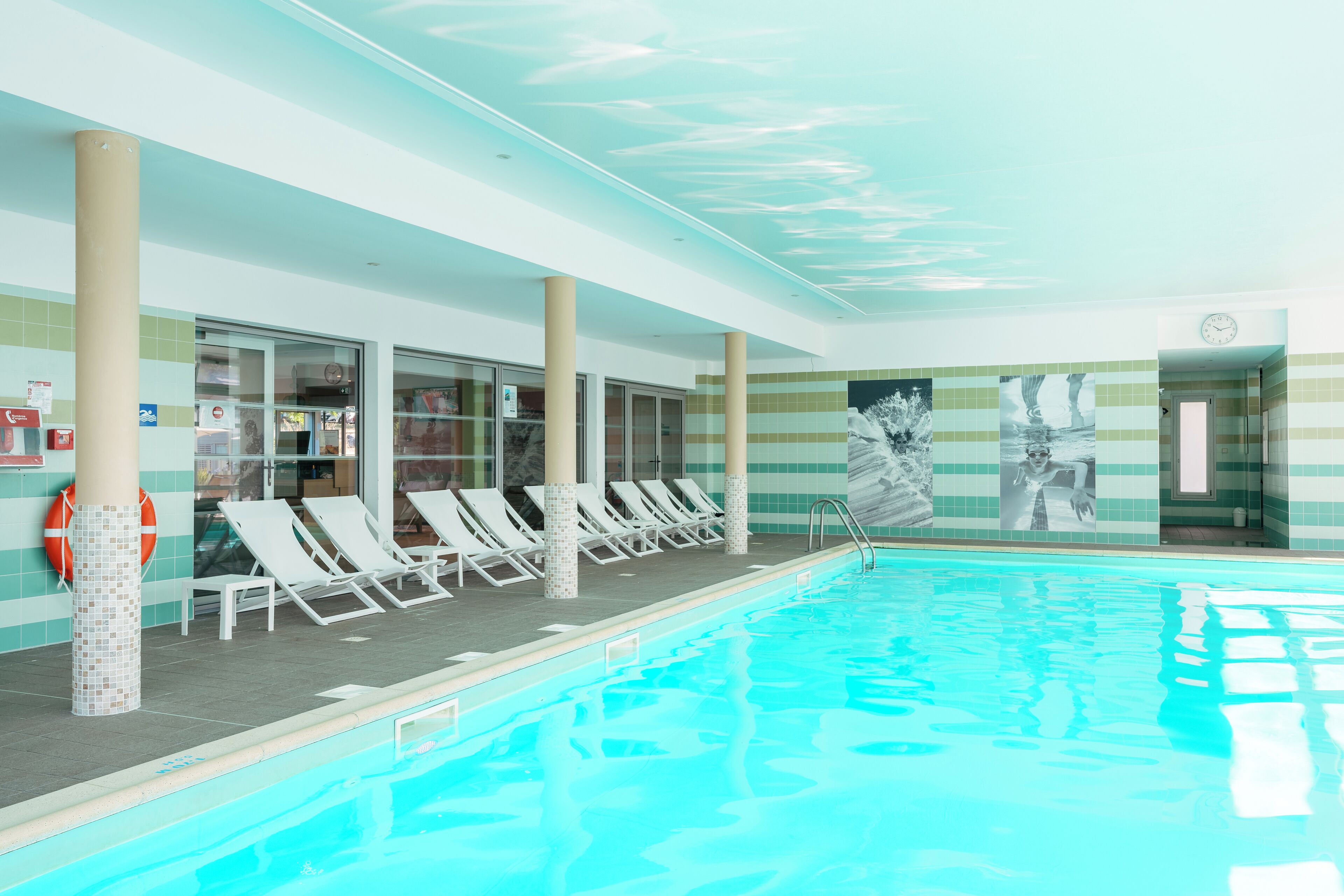 An indoor swimming pool with lounge chairs and a tiled wall with pictures.
