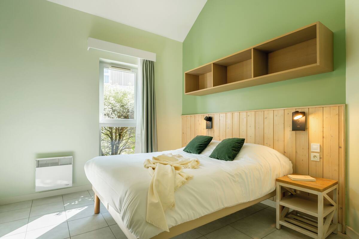 Une chambre à coucher comprenant un lit, une table de chevet, une étagère fixée au mur, une fenêtre avec des rideaux et un luminaire fixé au mur.