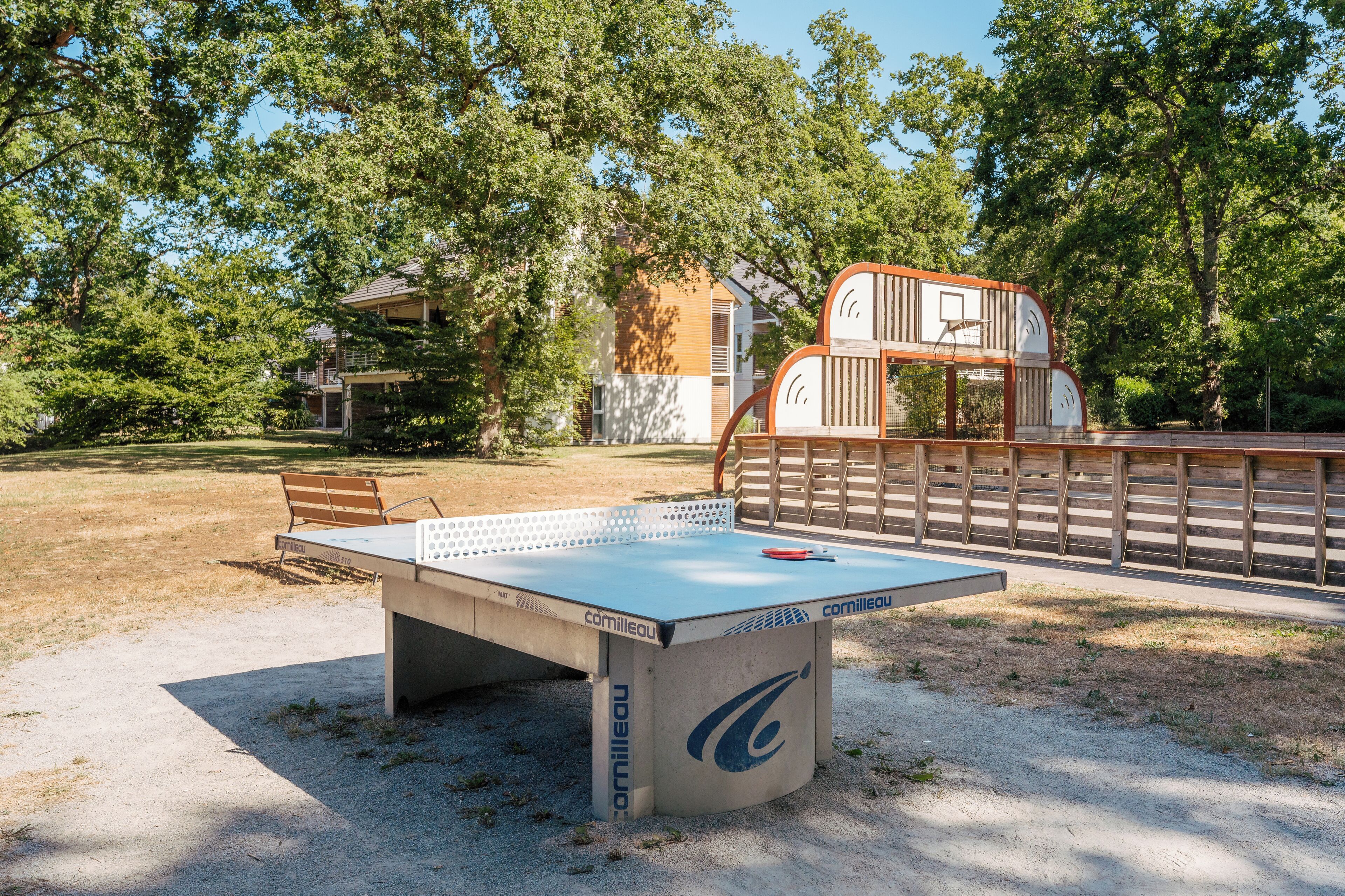Un tavolo da ping pong all'aperto con una panca di legno nelle vicinanze, circondato da alberi e un edificio sullo sfondo.