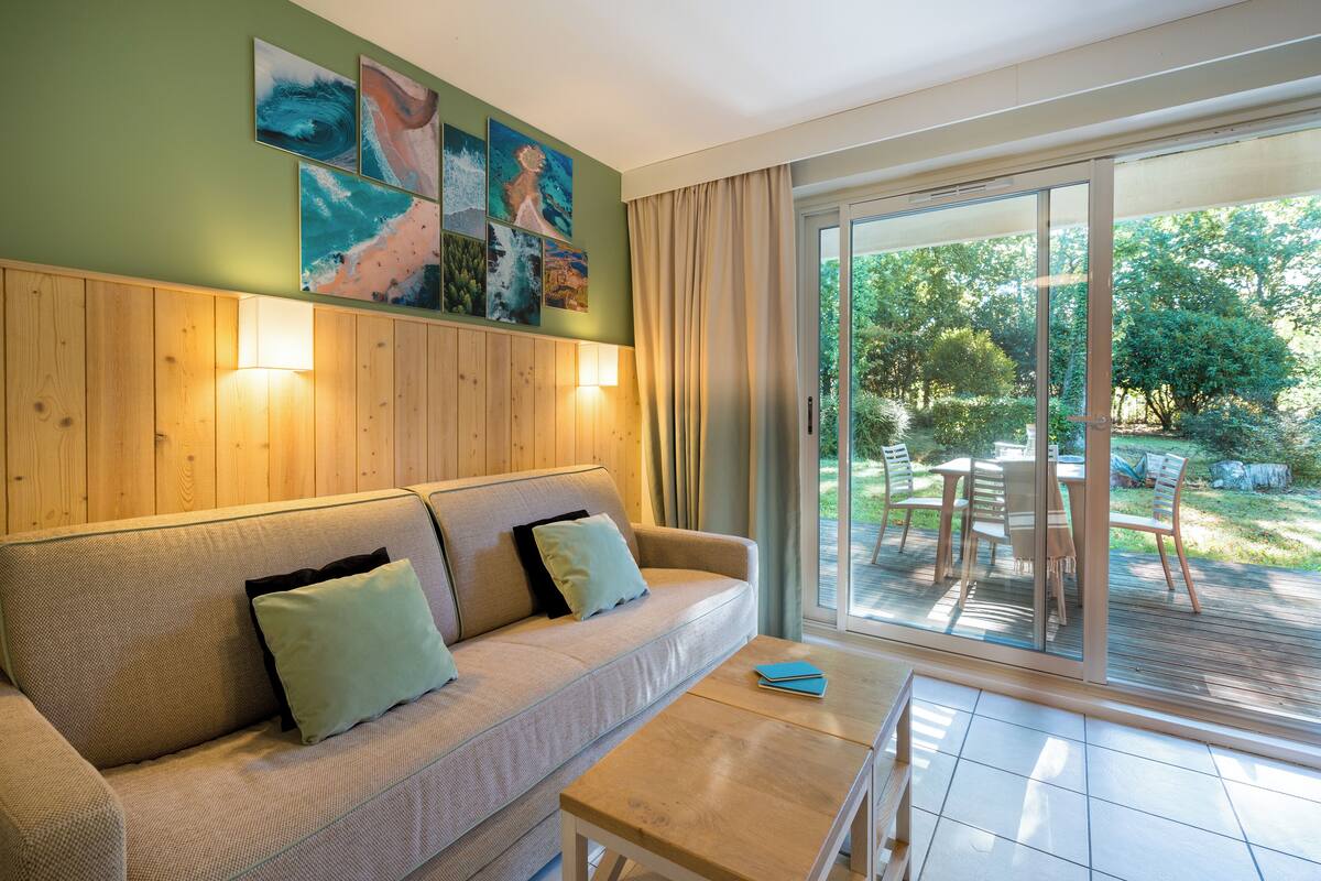 Un salon moderne avec un canapé, une table et une porte vitrée donnant sur un espace extérieur comprenant une terrasse et des chaises.