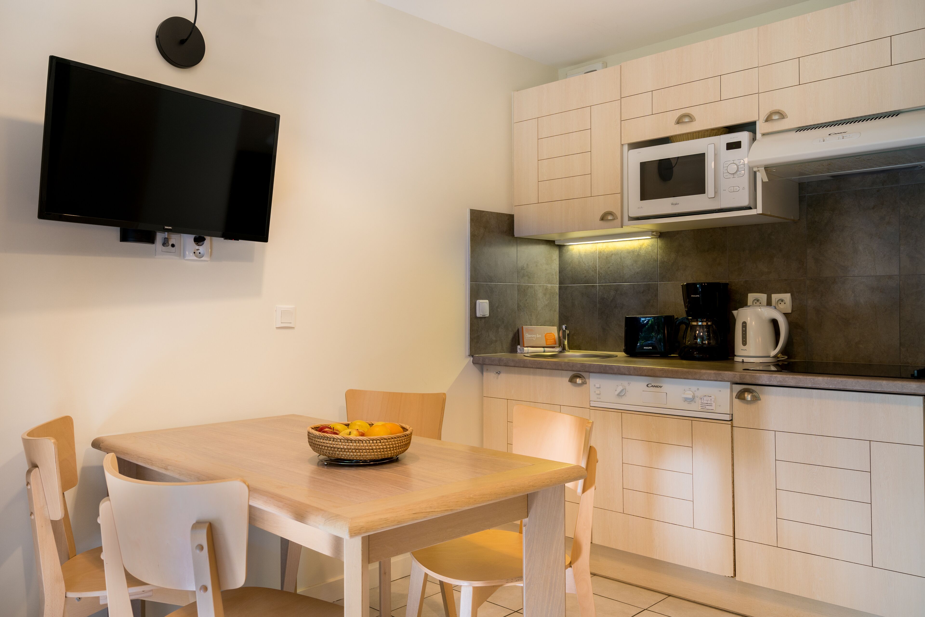 A compact kitchen with a flat-screen TV, a microwave, a coffee maker, and a bowl of fruit on the counter.