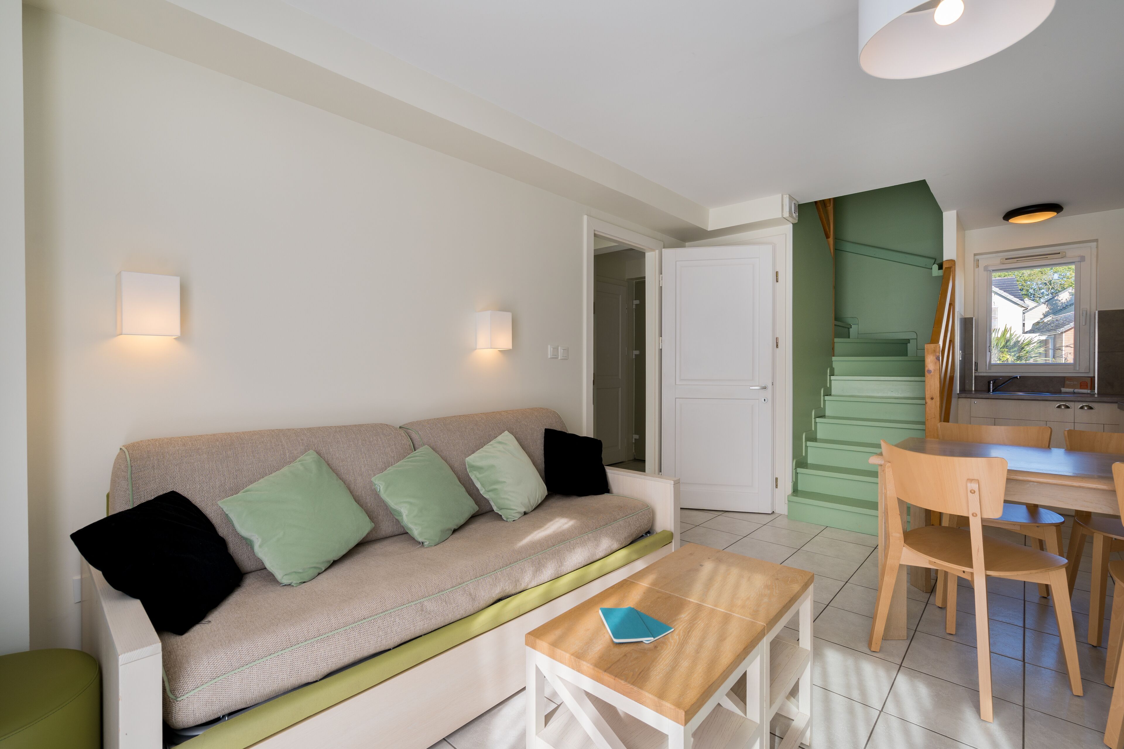 Ein modernes Wohnzimmer mit einem beigen Sofa, grünen Kissen, einem Holztisch und einer Küchenzeile mit einer grünen Treppe.