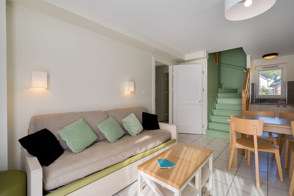 Una sala de estar moderna con un sofá color beige, cojines verdes, una mesa de centro de madera y una cocina con una escalera verde.