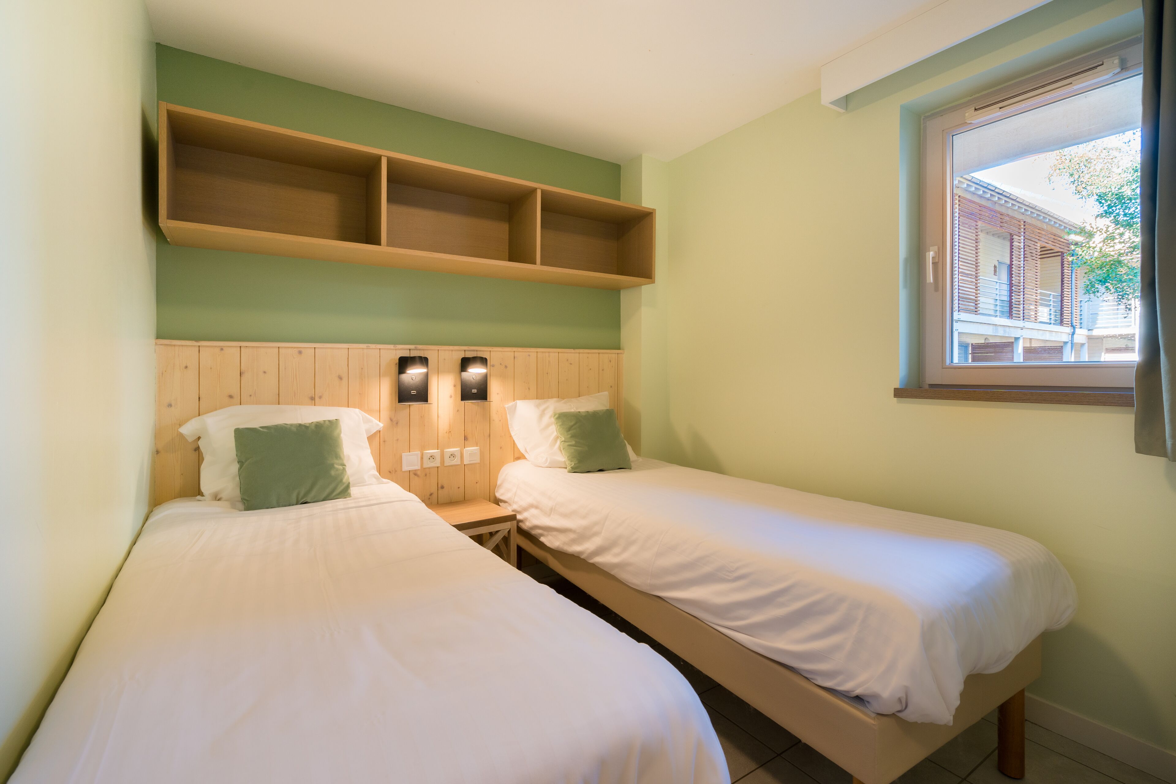 Ein Zimmer mit zwei Einzelbetten, einem Holz-Kopfstück, einem Regal über den Betten und einem Fenster mit Blick auf ein Gebäude.