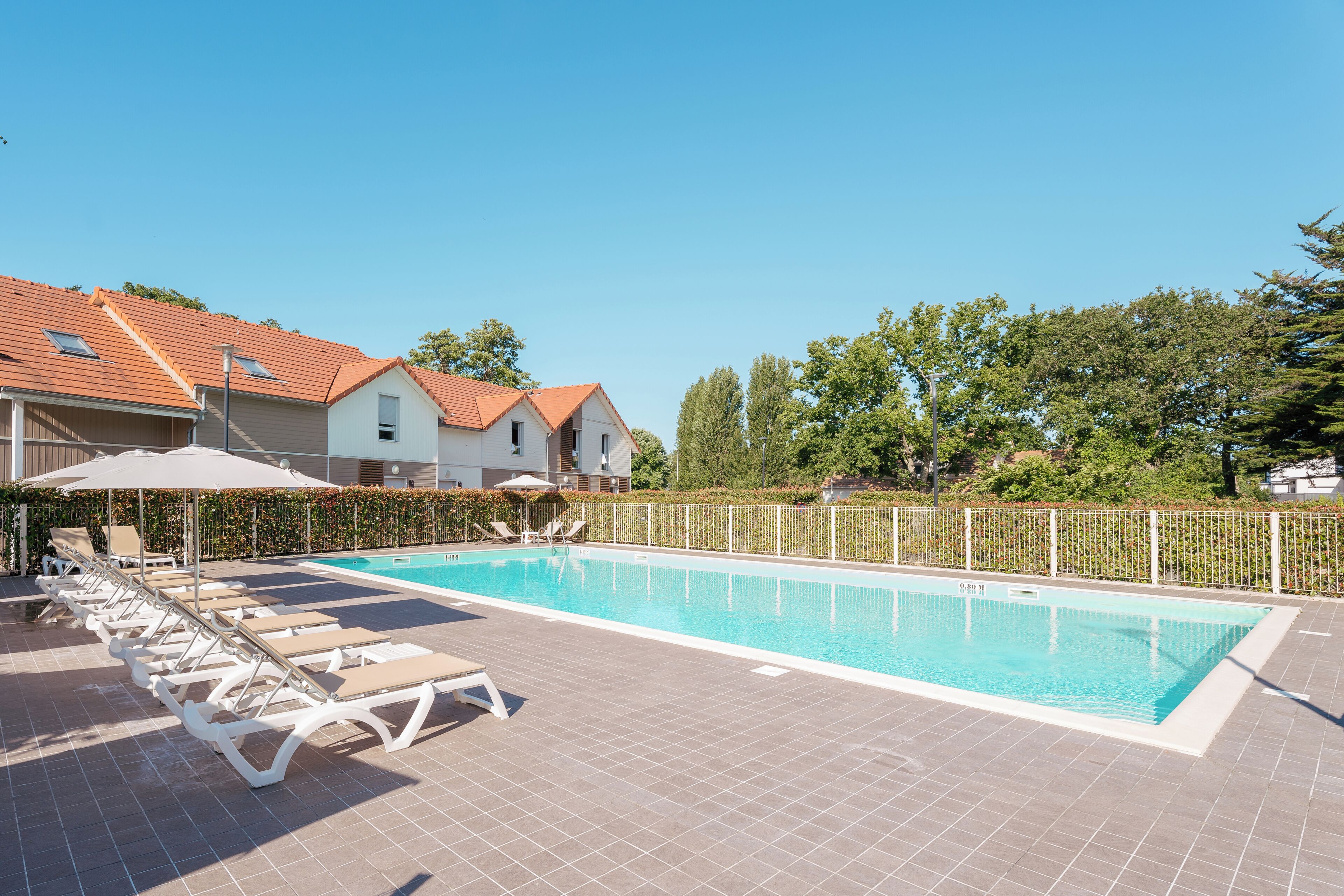 Uma piscina com espreguiçadeiras e guarda-sóis, cercada por uma grade e árvores.