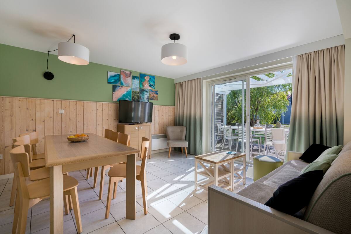 Un salon moderne avec une table à manger en bois, un canapé et un téléviseur. Une porte-fenêtre coulissante donne accès à un espace extérieur.