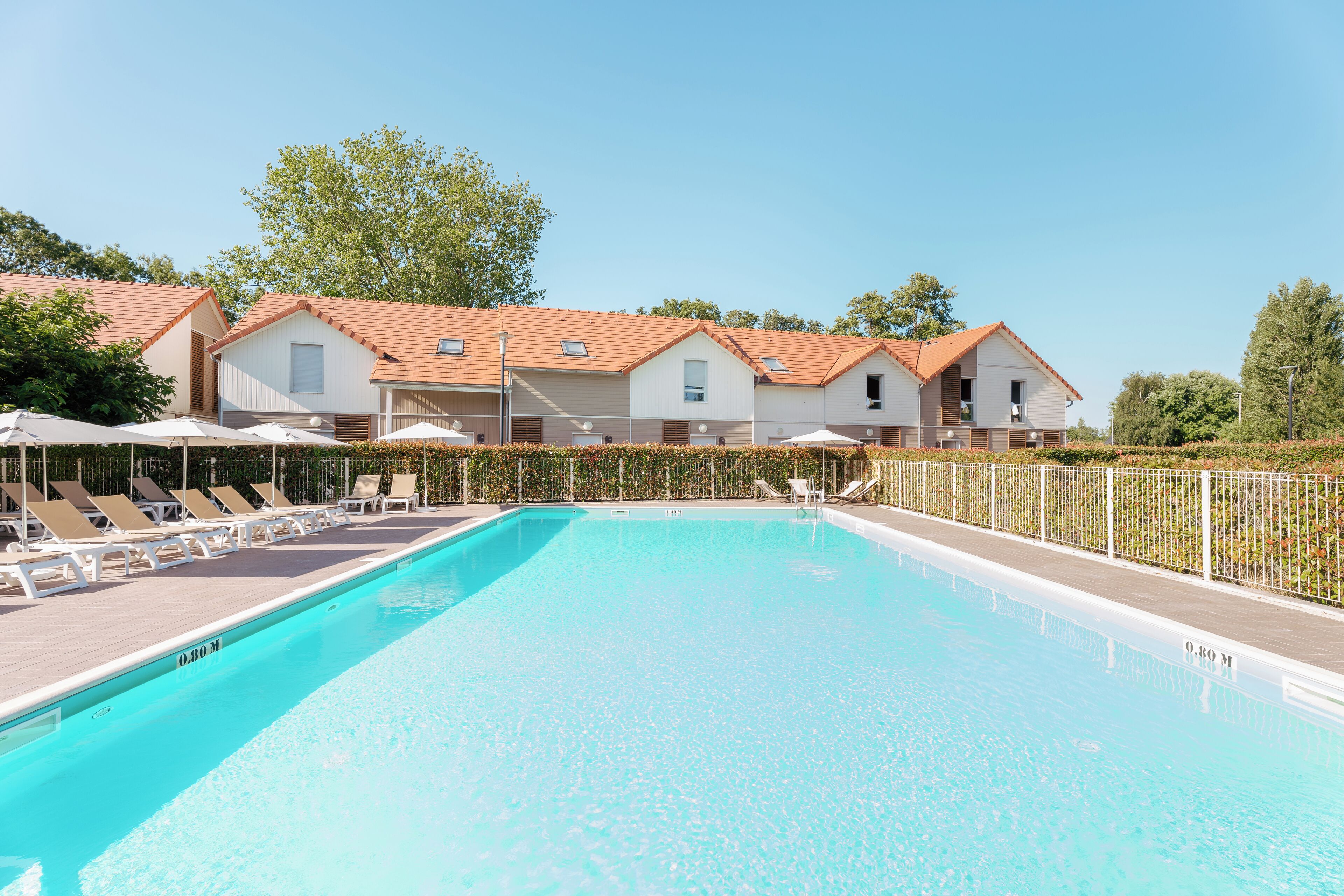 A swimming pool with lounge chairs and umbrellas, surrounded by a fence and buildings with red roofs.