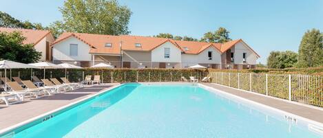 A swimming pool with lounge chairs and umbrellas, surrounded by a fence and buildings with red roofs.