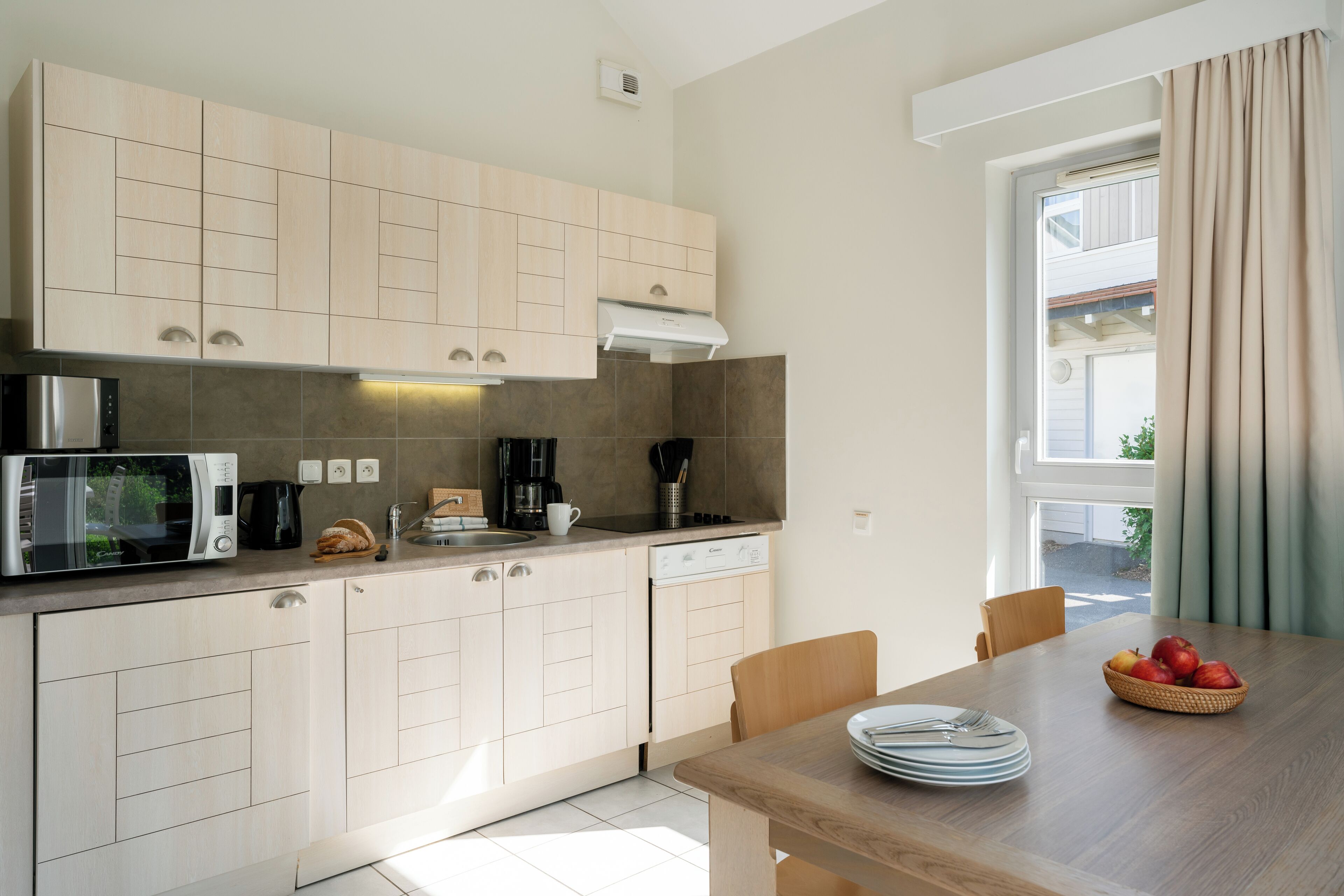 A modern kitchen with light wood cabinets, a microwave, coffee maker, and a dishwasher. There is a dining table with plates and a fruit basket.