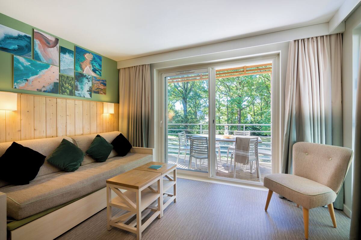 Une pièce moderne comprenant un canapé, un fauteuil et une table. Elle est dotée d’une grande fenêtre donnant sur des arbres et d’un balcon.