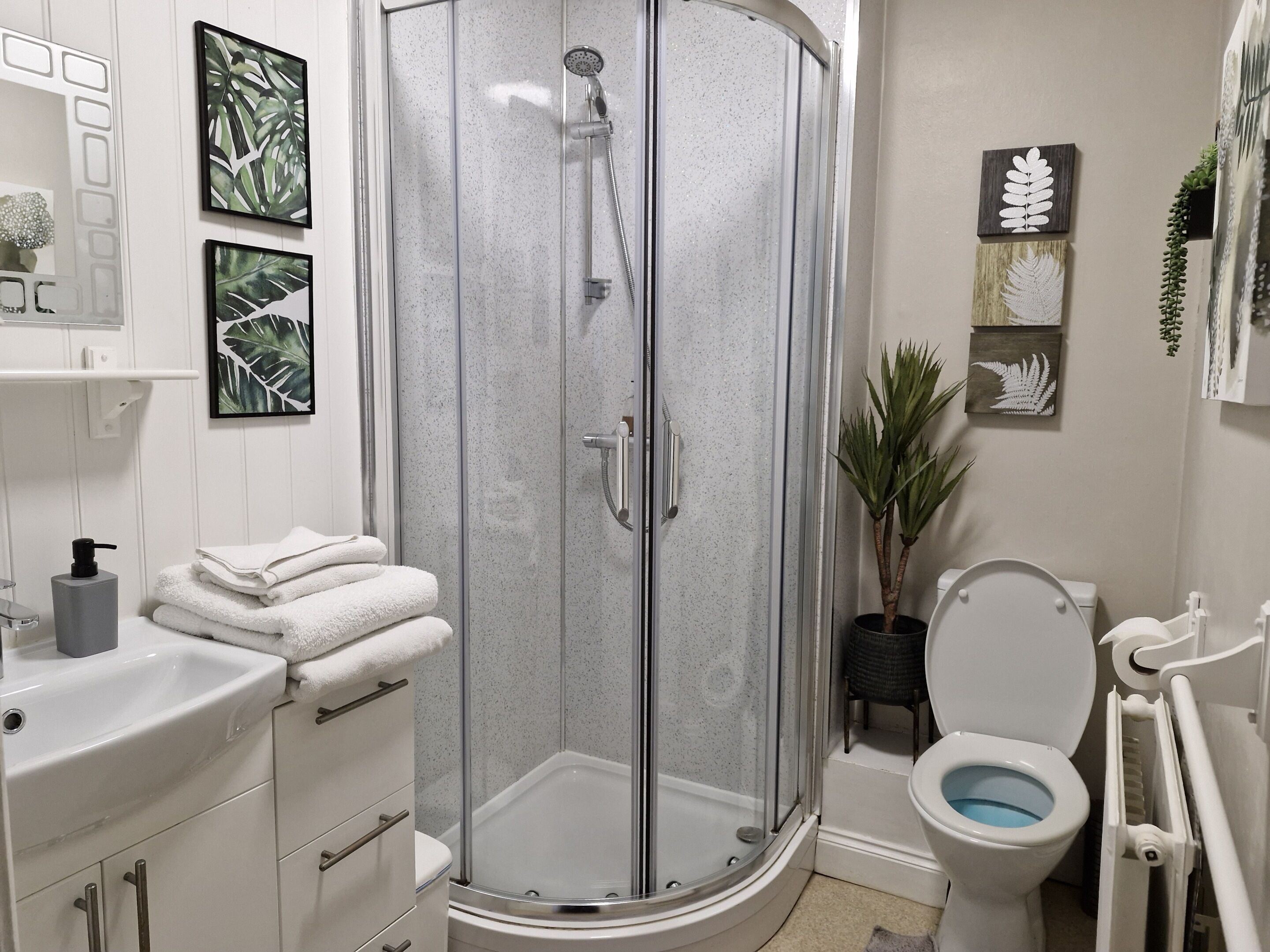 Een moderne badkamer met een douche met glazen wand, witte sanitair en decoratieve bladkunst aan de muur.