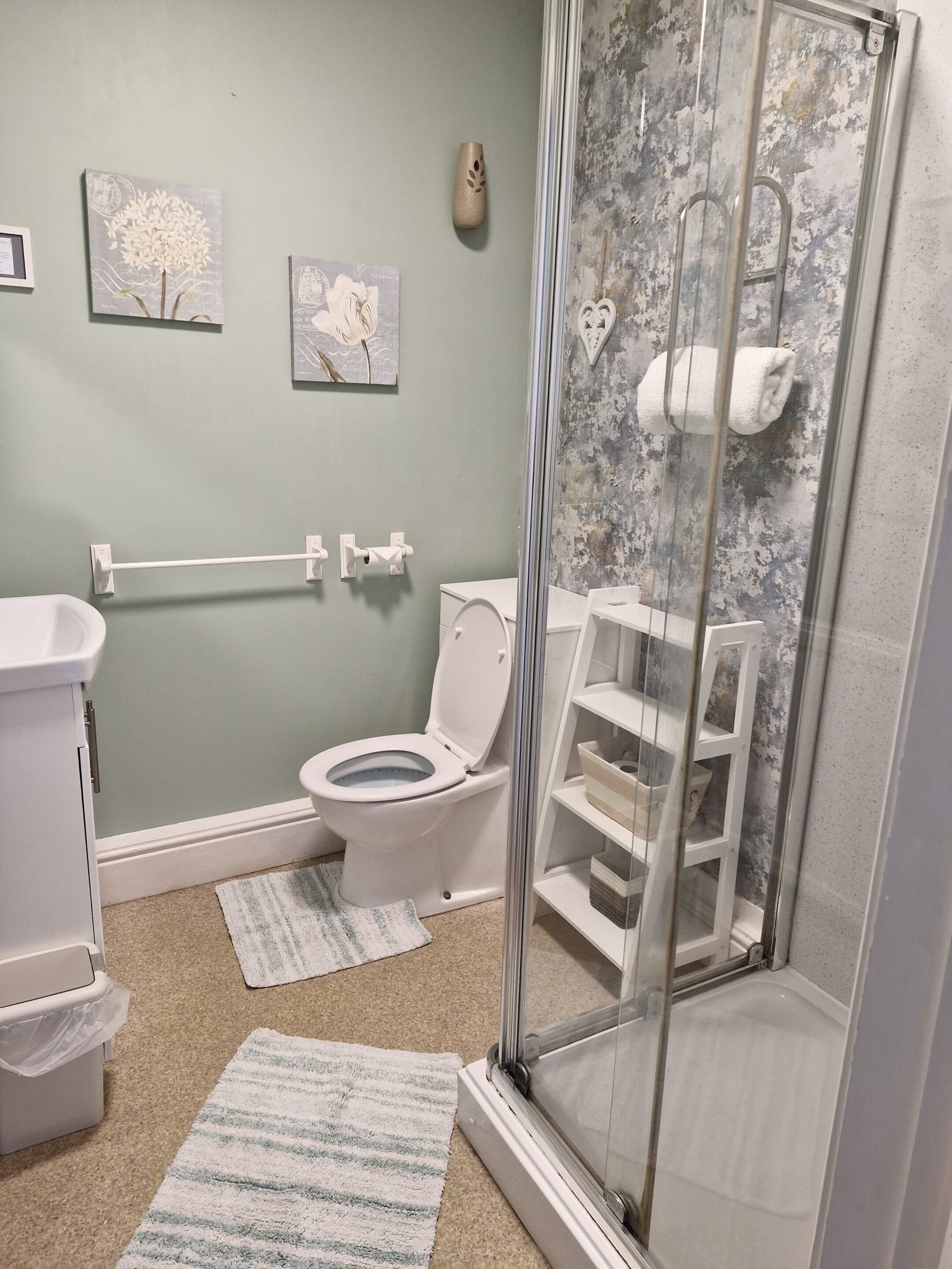 Et badeværelse med toilet, håndvask, bruser med glasdør og to håndklæder på gulvet.