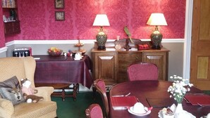 A dining room with a wooden table set for four, two lamps, a red patterned wallpaper, and a wooden cabinet.