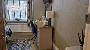 A hotel room with a desk, a chair, a TV mounted on the wall, a bedside table with a lamp, a bedside plant, and a window with curtains.