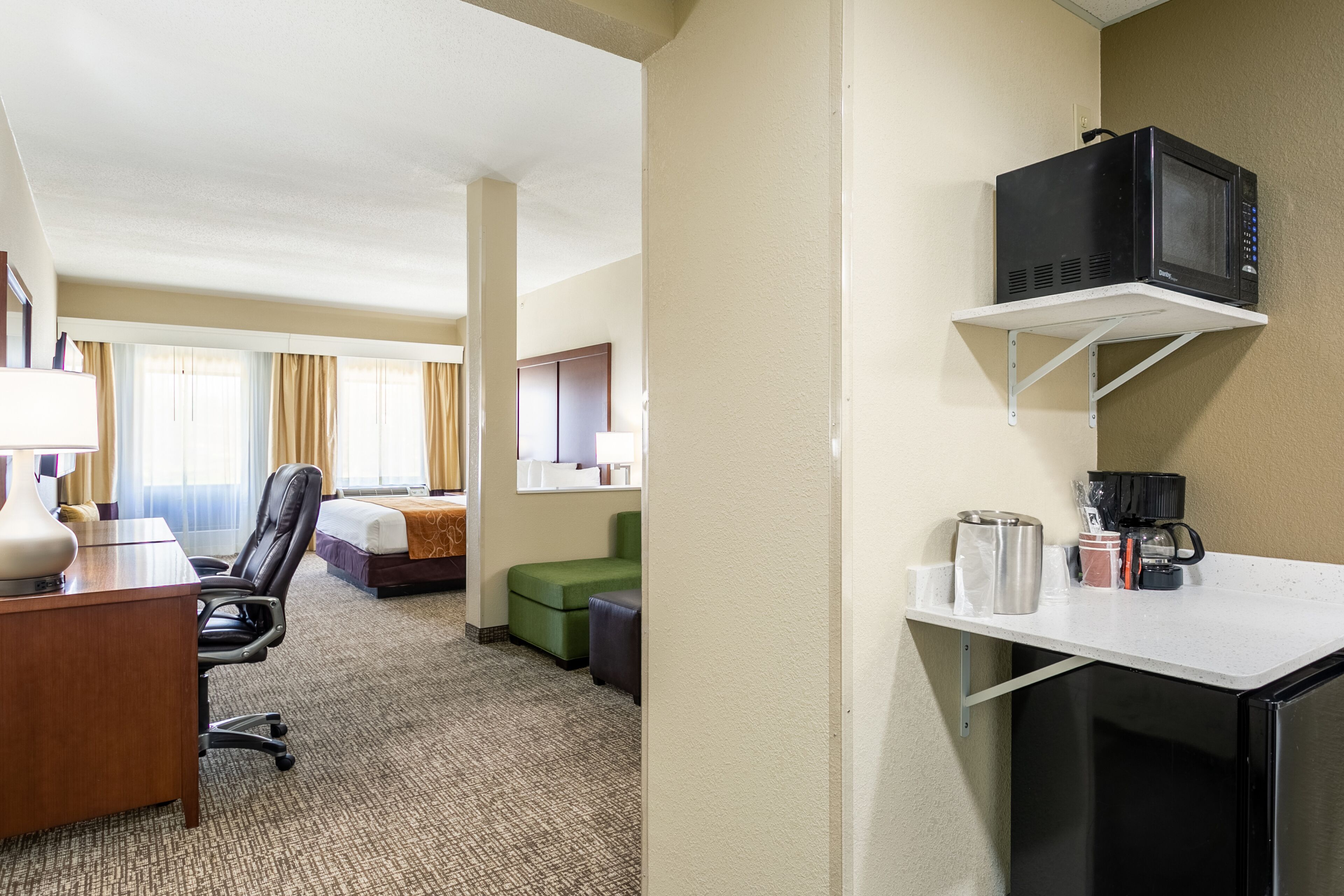 A hotel room with a desk, chair, bed, and a kitchenette area with a microwave, coffee maker, and storage containers.