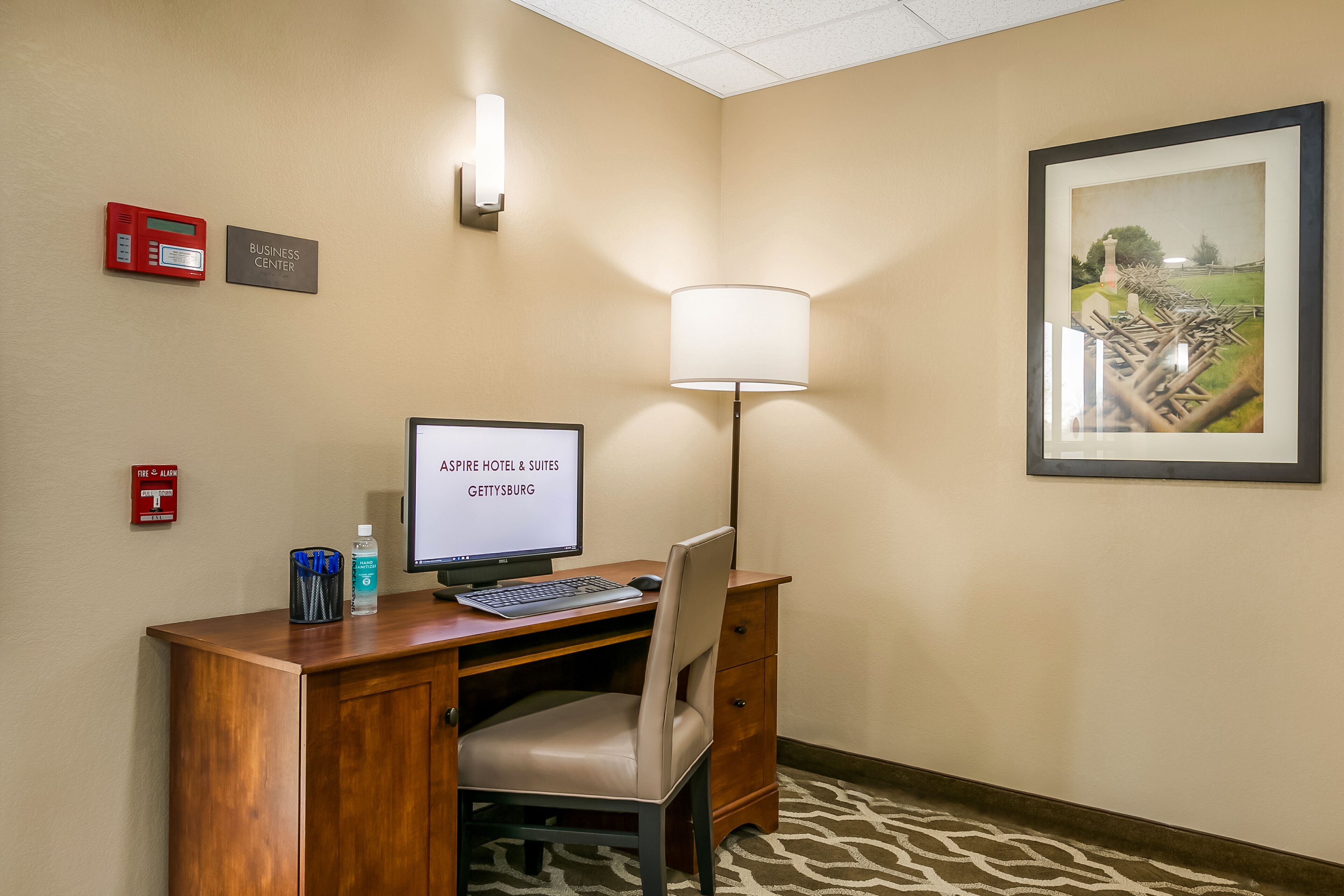 A hotel room with a desk, computer, chair, lamp, framed picture, and wall-mounted signs including a fire alarm and business center.
