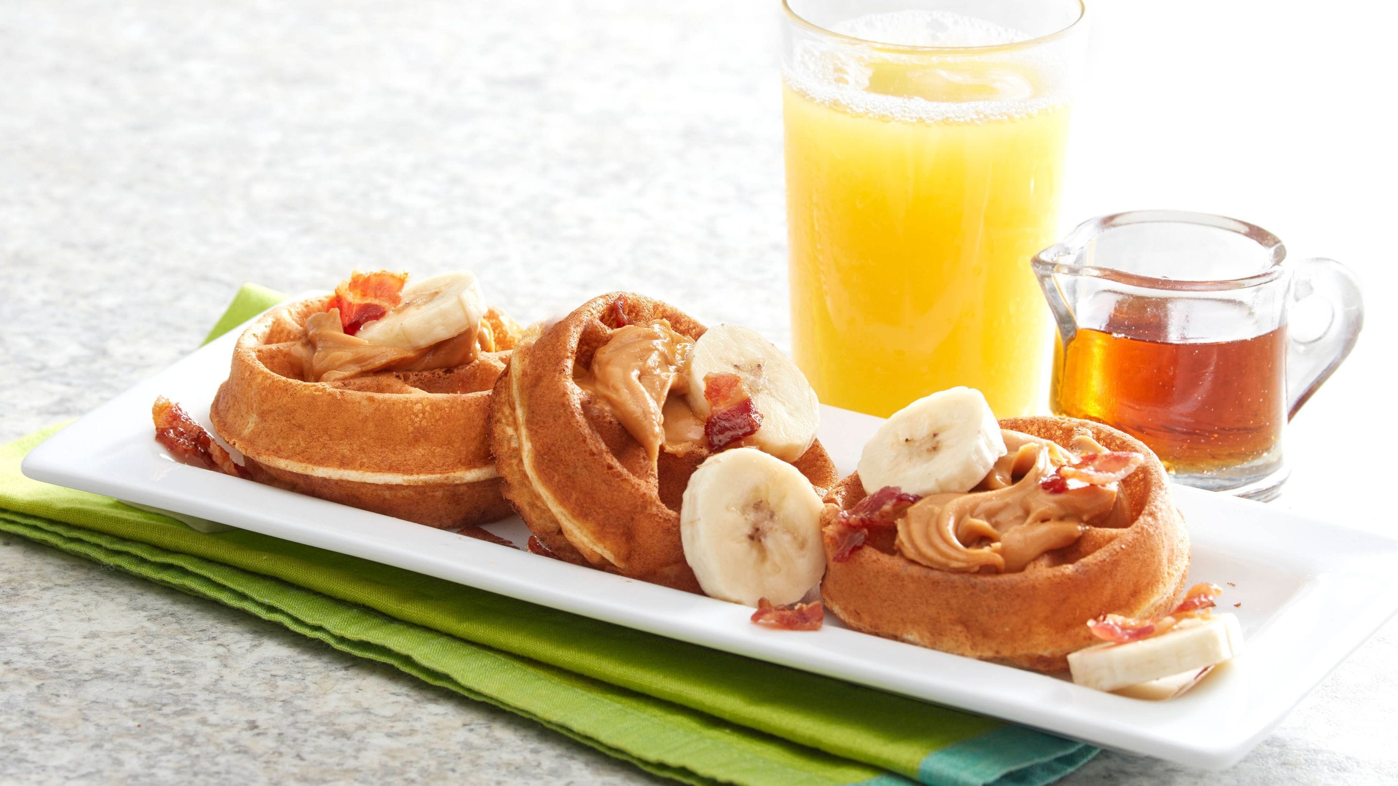 A breakfast plate with pancakes, bananas, and bacon, accompanied by a glass of orange juice and a mug of tea.