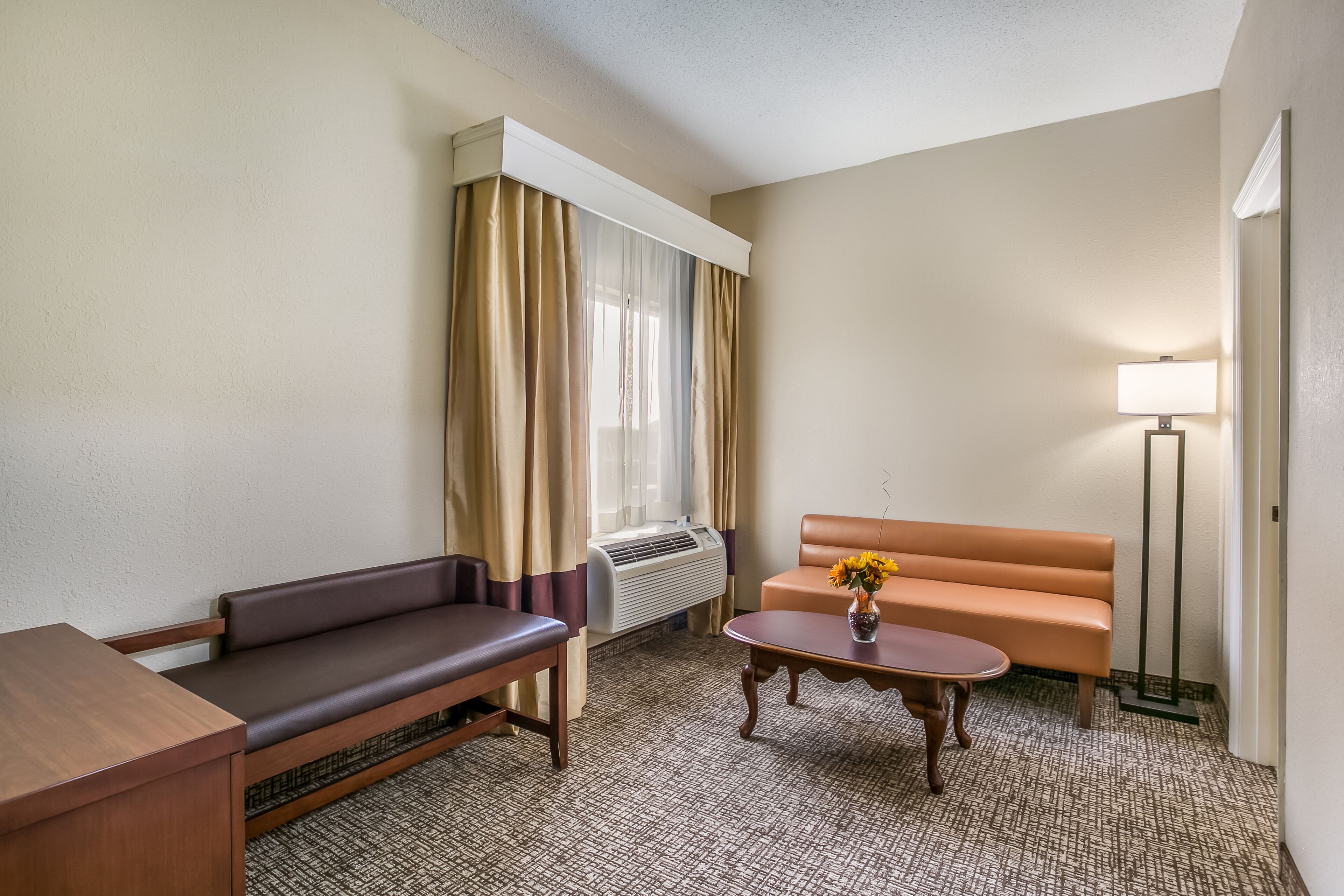 Ein Hotelzimmer mit einer Couch, einem kleinen Tisch mit Blumenvase, einer Lampe und einer Klimaanlage.