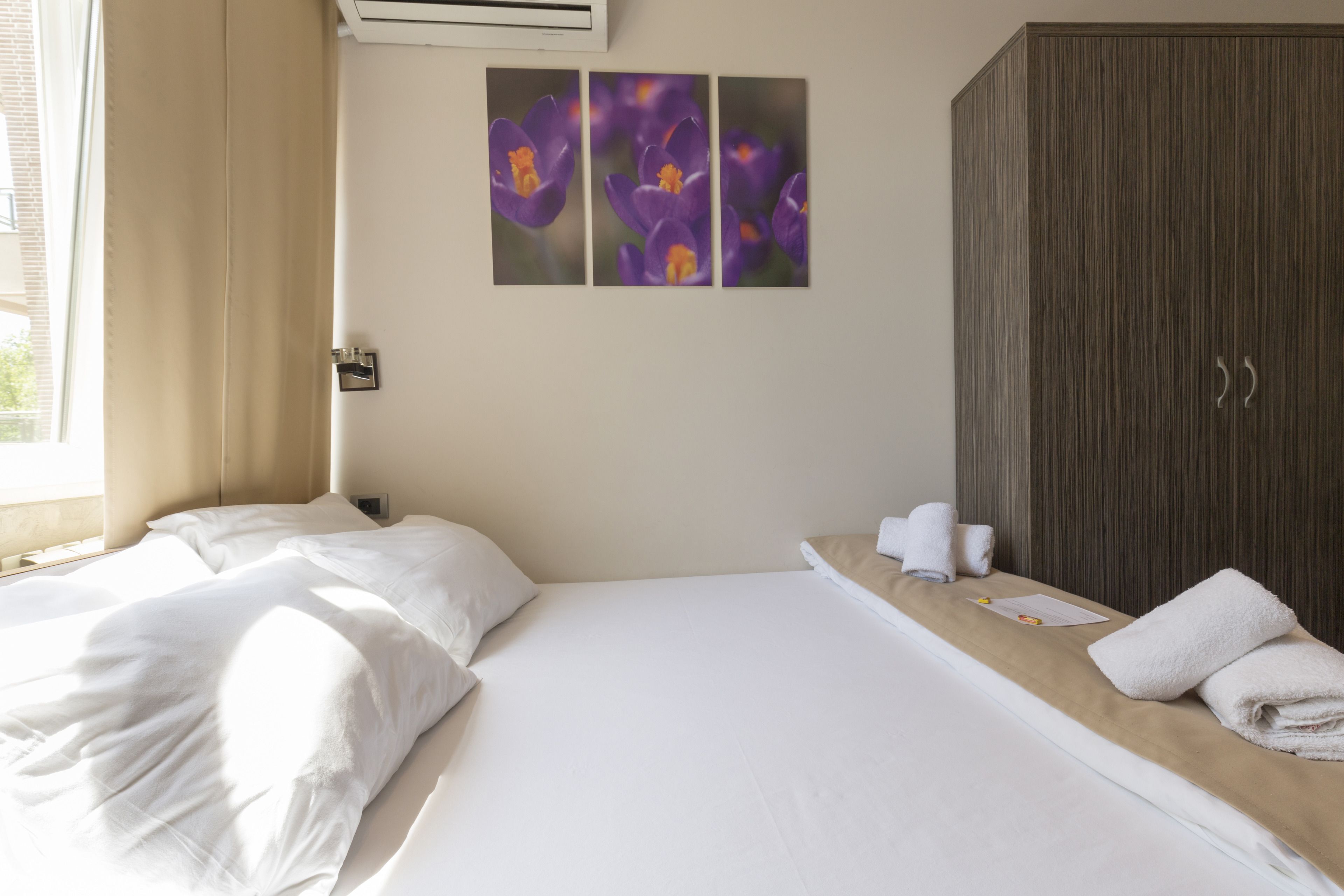 Una habitación de hotel con cama, mesita de noche, armario y ventana con cortinas.