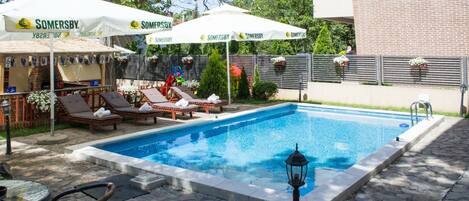 Relax by the rectangular pool surrounded by lounge chairs and umbrellas, with a brick building in the background.