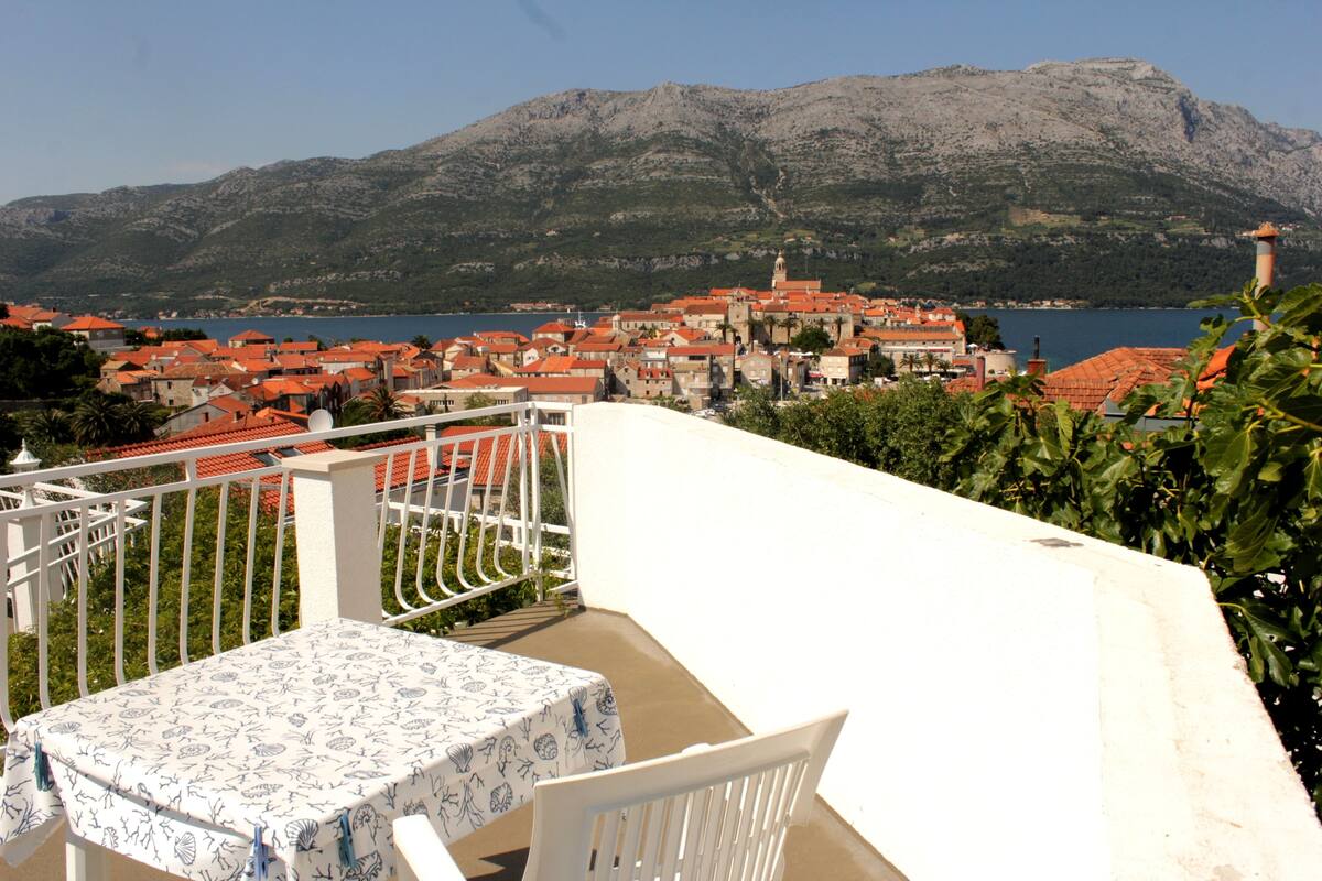 A balcony with a table and chair, overlooking a coastal town with red-roofed buildings and a mountain in the background.