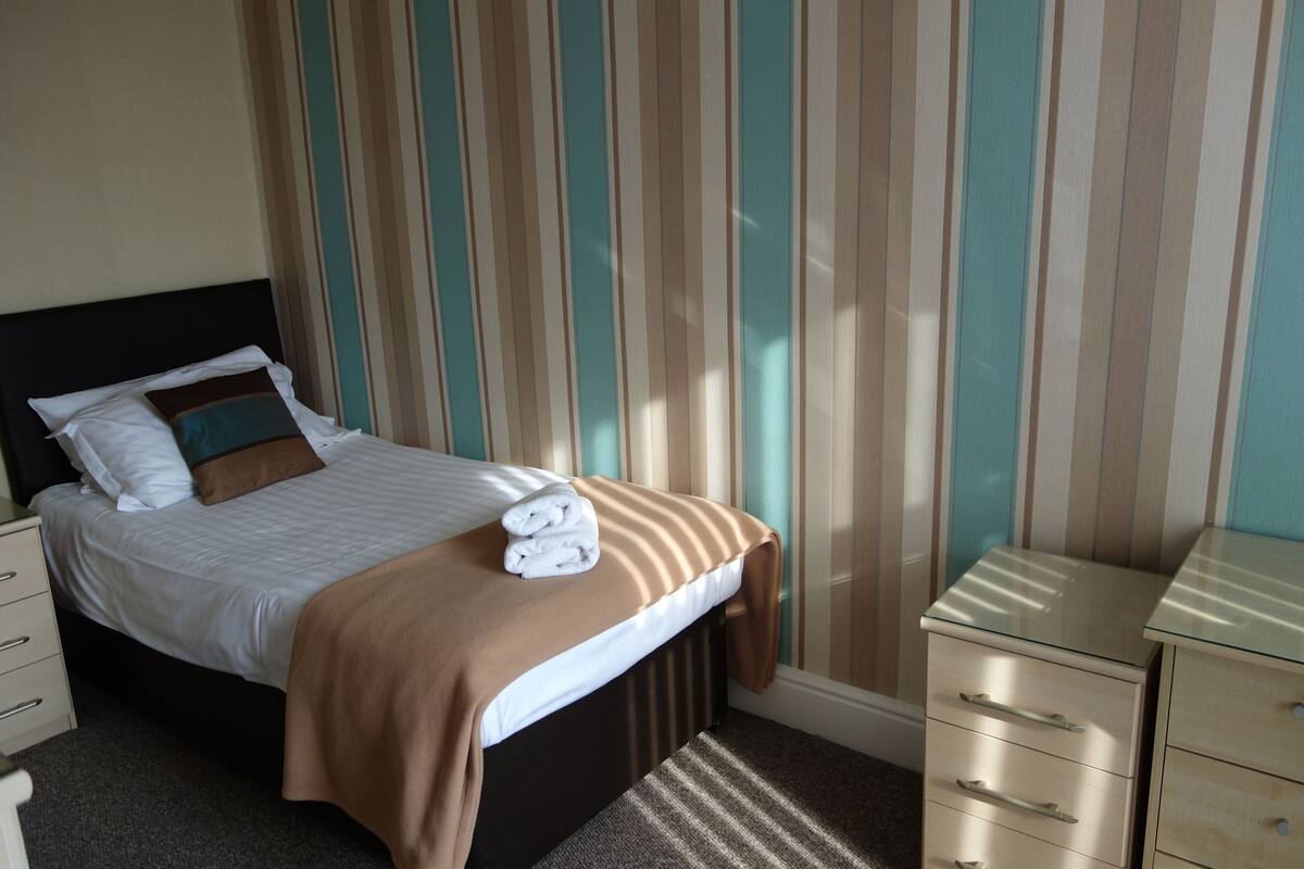 A neatly made bed with a brown blanket and two folded towels, a bedside table with drawers, and a striped curtain in the background.