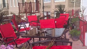 Terraza al aire libre con sillas y sombrilla rojos, rodeada de plantas y flores en macetas.