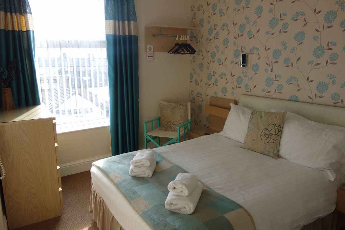 A hotel room with a bed, a desk, and a chair. There are towels on the bed and a window with curtains.