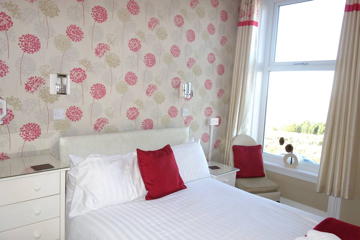 A hotel room with a bed, bedside tables, a nightstand, a window with curtains, and a wall with floral wallpaper.