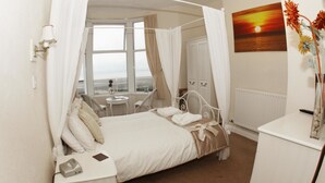 Un dormitorio con cama con dosel, una mesita de noche con lámpara, vista al mar a través de una ventana y un cuadro de un atardecer.