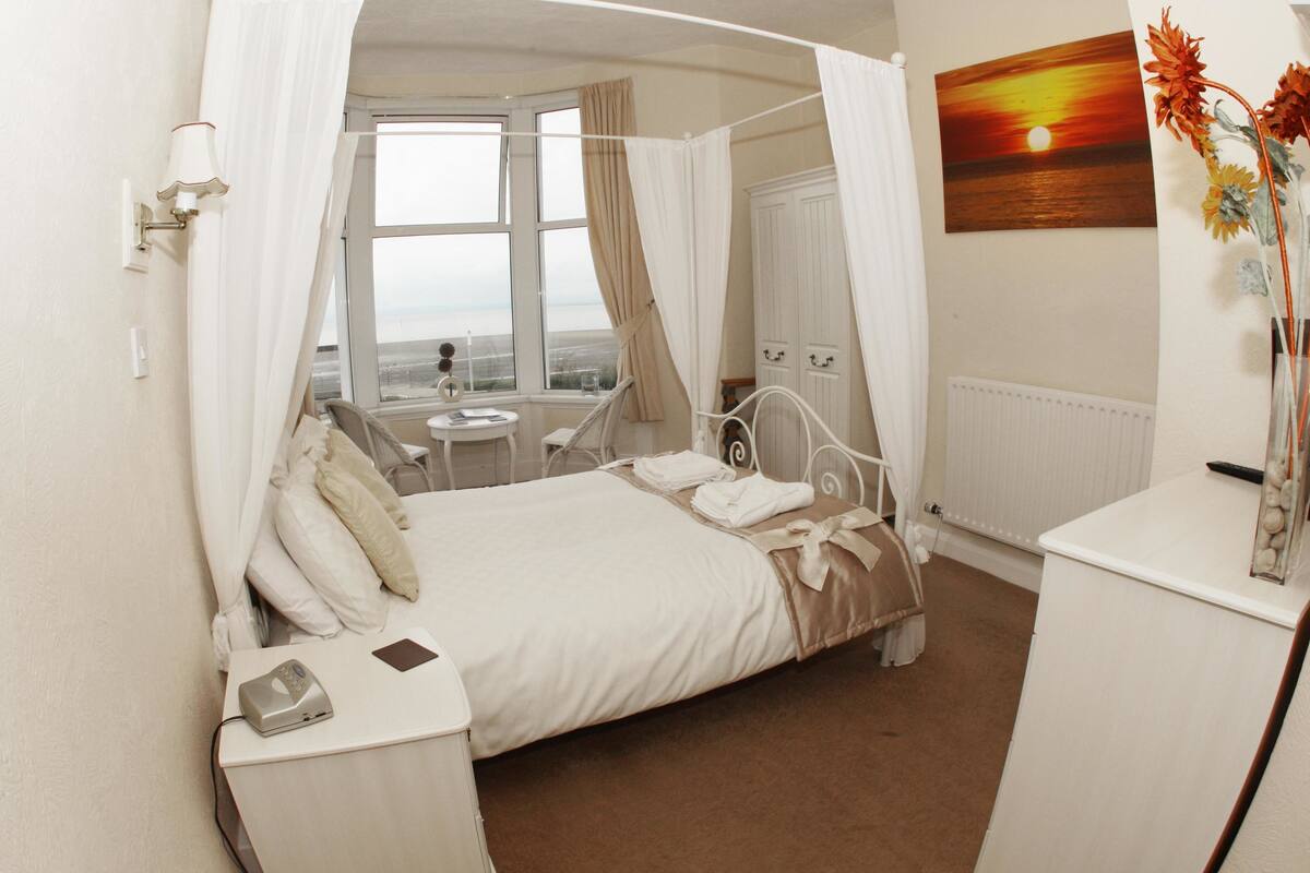 A bedroom with a four-poster bed, a bedside table with a lamp, a view of the sea through a window, and a painting of a sunset.