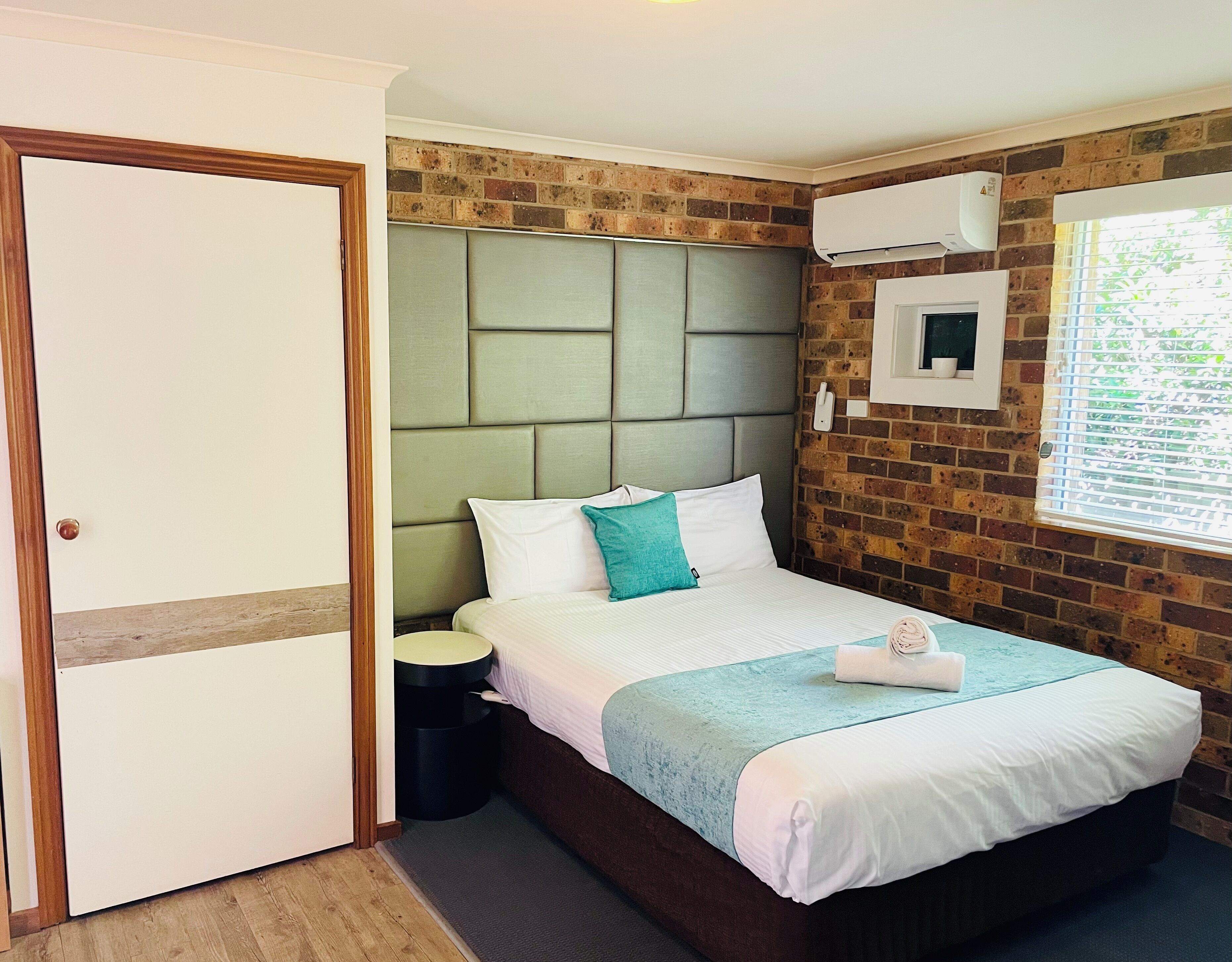 A hotel room with a brick wall, a bed with white bedding and a teal pillow, a bedside table, a small round nightstand, a white air conditioning unit, and a window with blinds.
