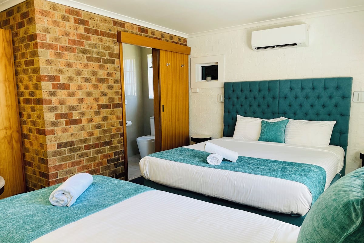 Habitación de hotel con dos camas, una pared de ladrillo, un cabecero color verde azulado y un equipo de aire acondicionado.