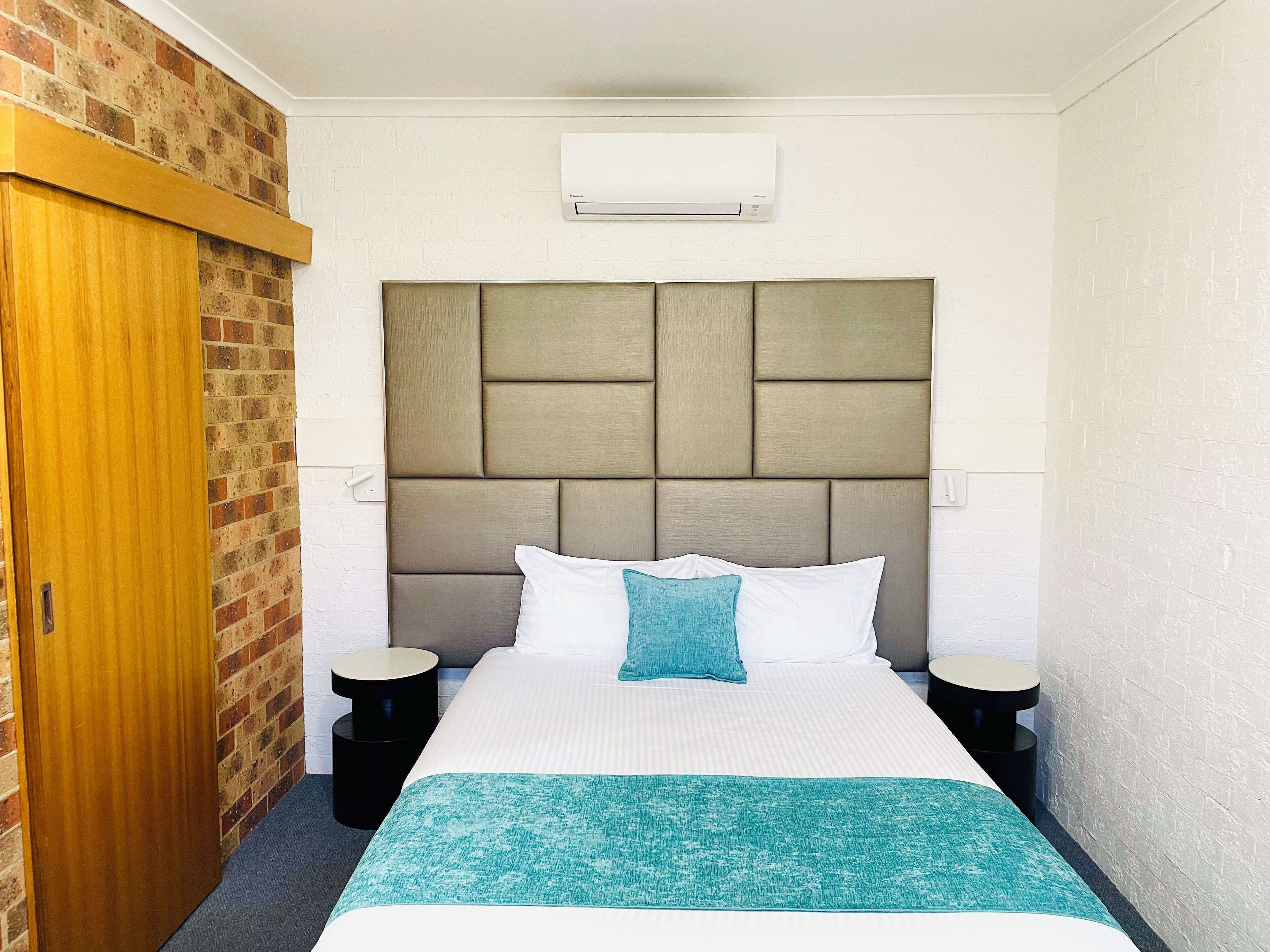 Habitación de hotel con pared de ladrillo decorativa, puerta de madera, cama grande con cabecera acolchada, dos mesitas de noche y un equipo de aire acondicionado en la pared.
