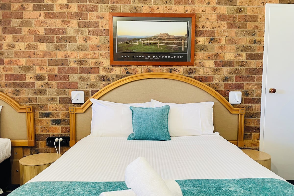 Habitación de hotel con pared de ladrillo, un cuadro enmarcado de un paisaje, una cama con ropa de cama blanca y turquesa, y una toalla enrollada sobre la cama.