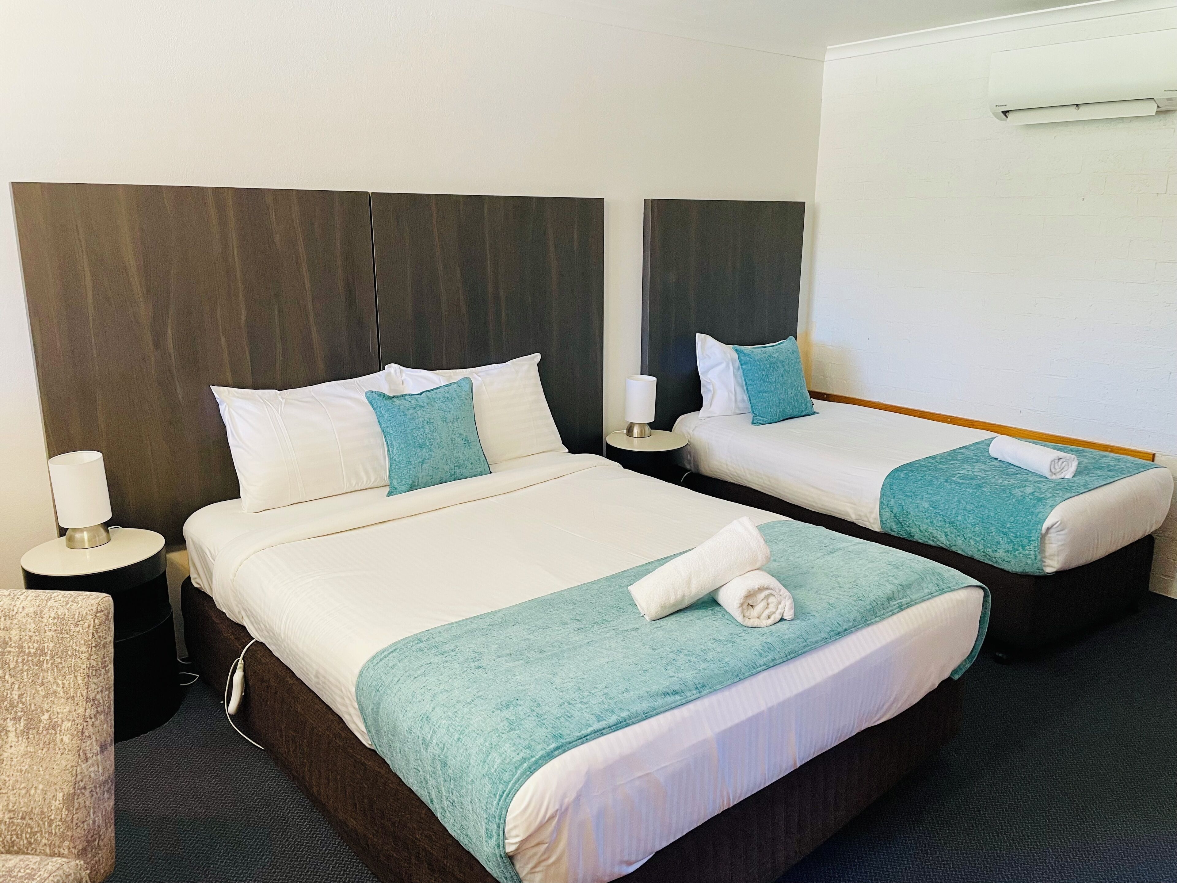 Hotellihuone, jossa on kaksi sänkyä, yöpöytä lampun kanssa ja seinälle asennettu ilmastointilaite.