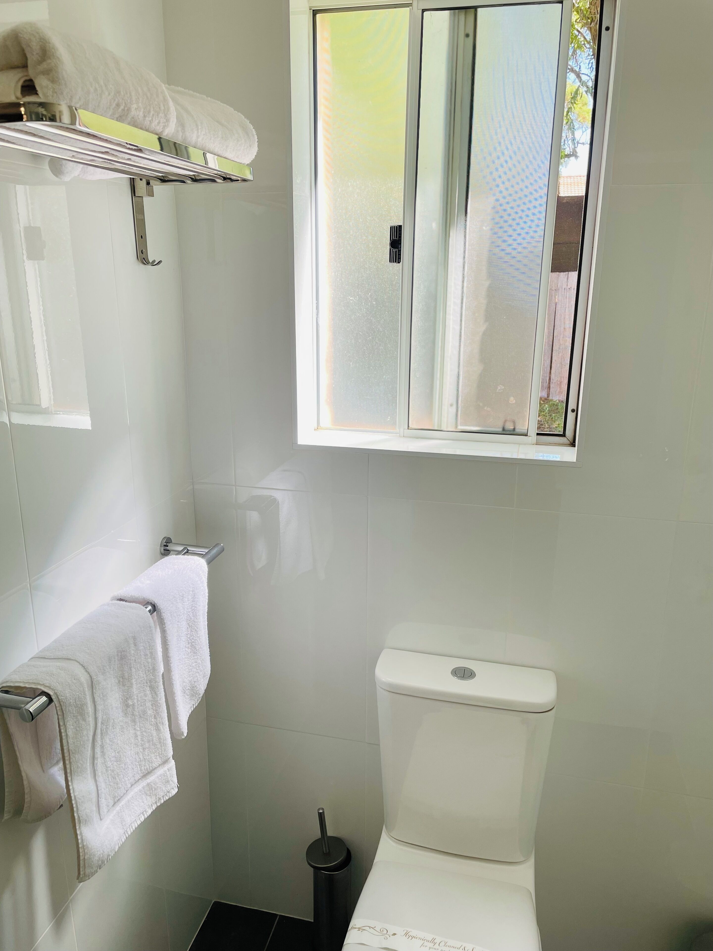 A bathroom with a toilet, a towel rack, a window, and a trash bin.