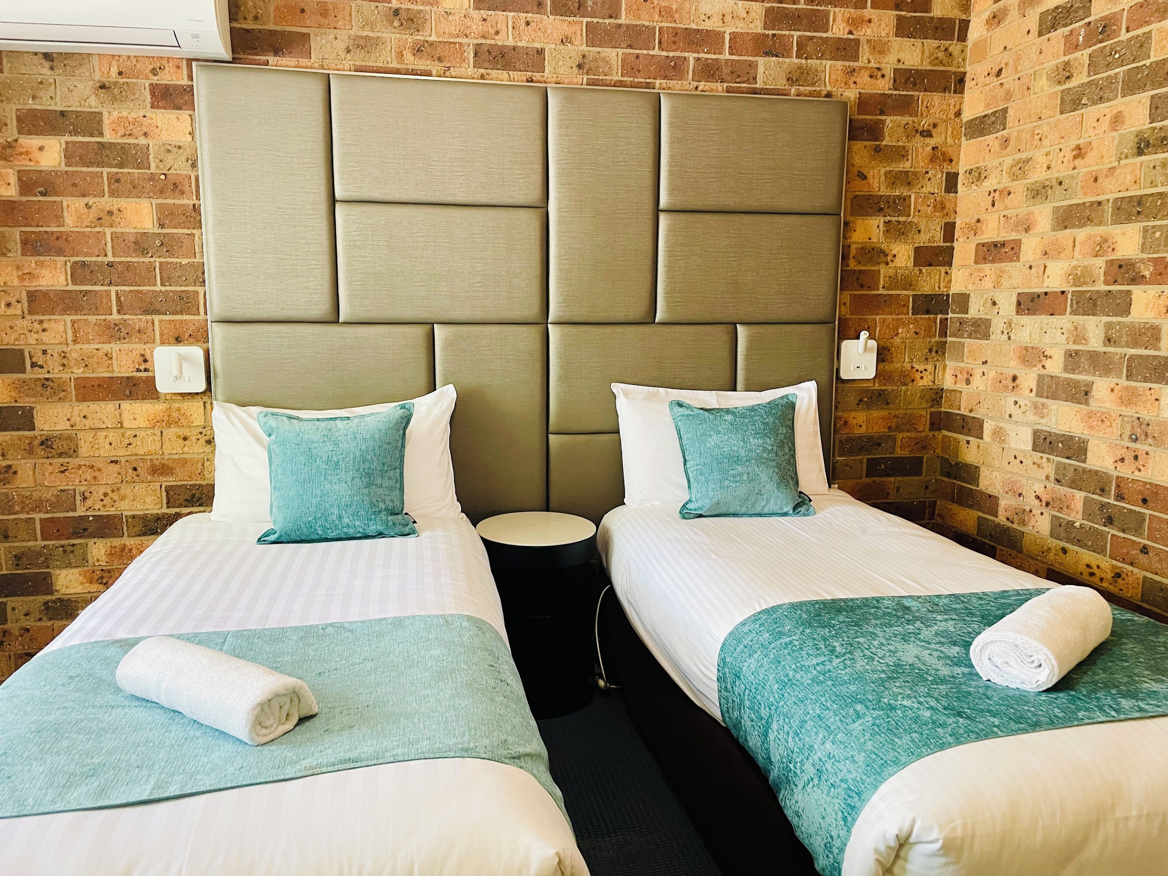 A hotel room with two beds, a brick wall, a modern headboard, and a small round table.