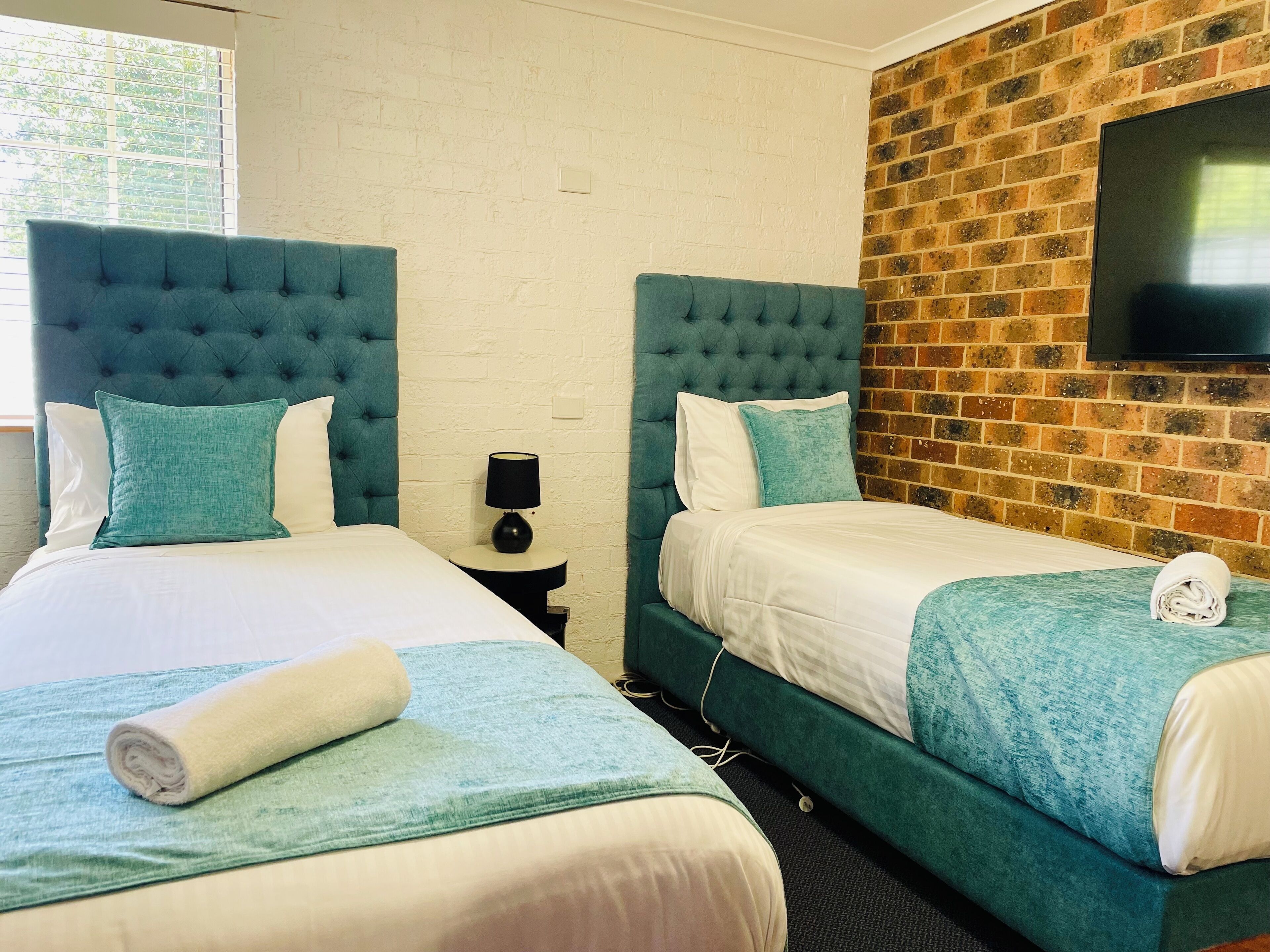 A hotel room with two beds, each with a tufted headboard, a flat-screen TV mounted on a brick wall, and a small bedside table with a lamp.