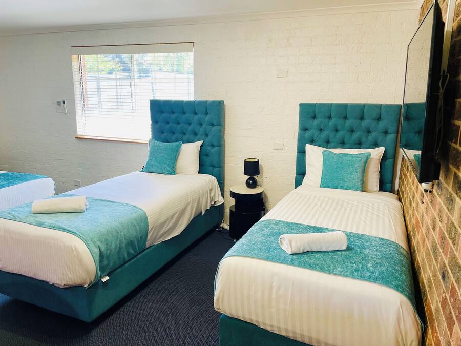 Habitación de hotel con dos camas, cada una con cabecero acolchado, una mesita de noche con lámpara y una ventana con persianas.