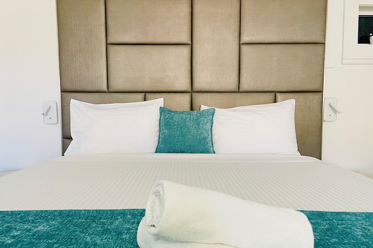 Una cama con cabecera, almohadas blancas, un cojín decorativo azul aguamarina y una toalla blanca enrollada sobre un edredón azul aguamarina.