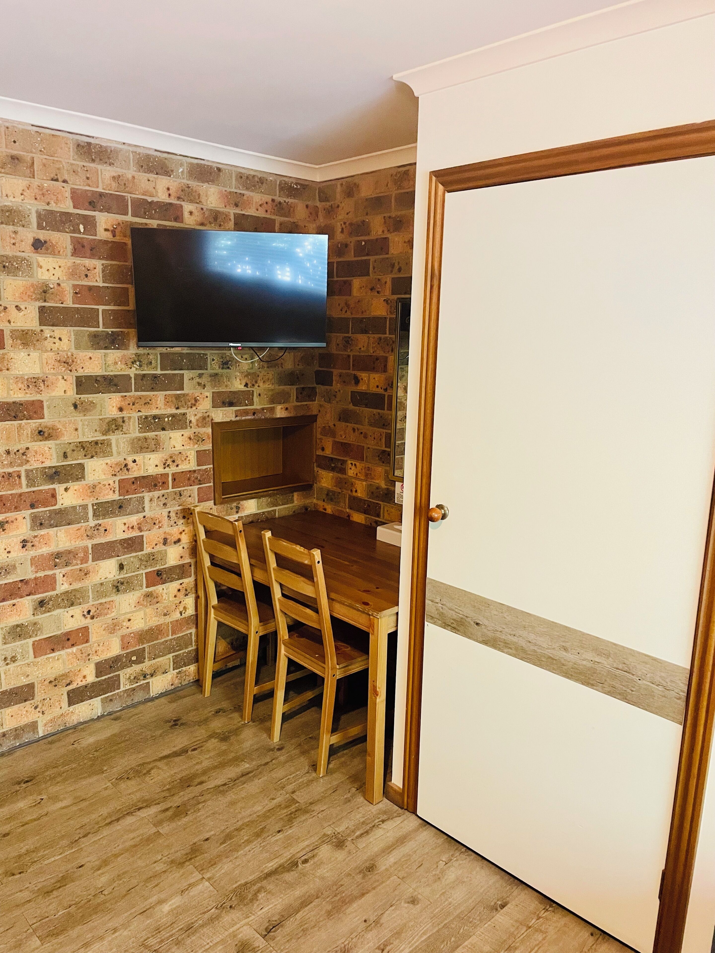 Een kamer met een bakstenen muur, een flatscreen televisie boven een houten tafel met stoelen, en een witte deur.