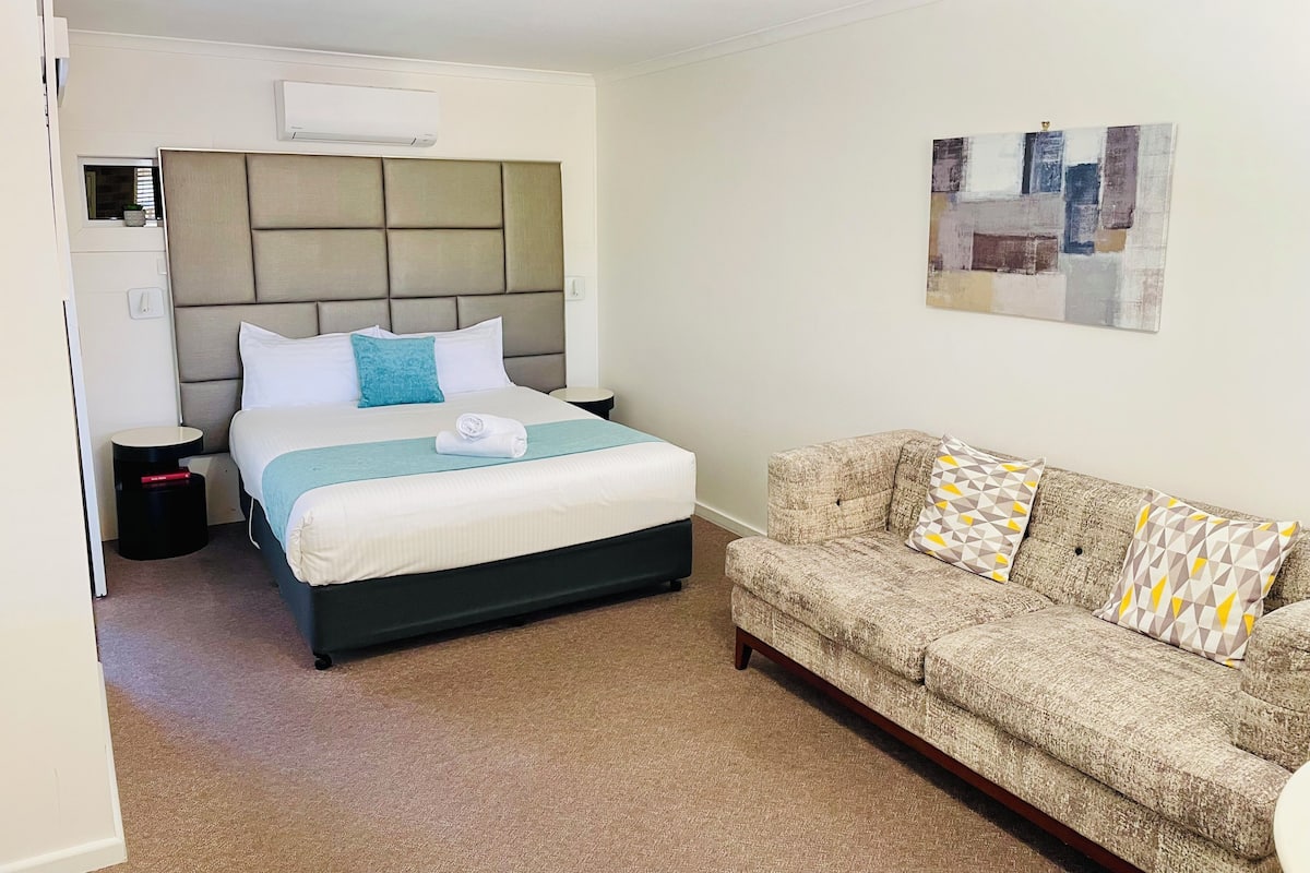 Una habitación de hotel con cama, sofá, aire acondicionado instalado en la pared, una mesita redonda y un cuadro.