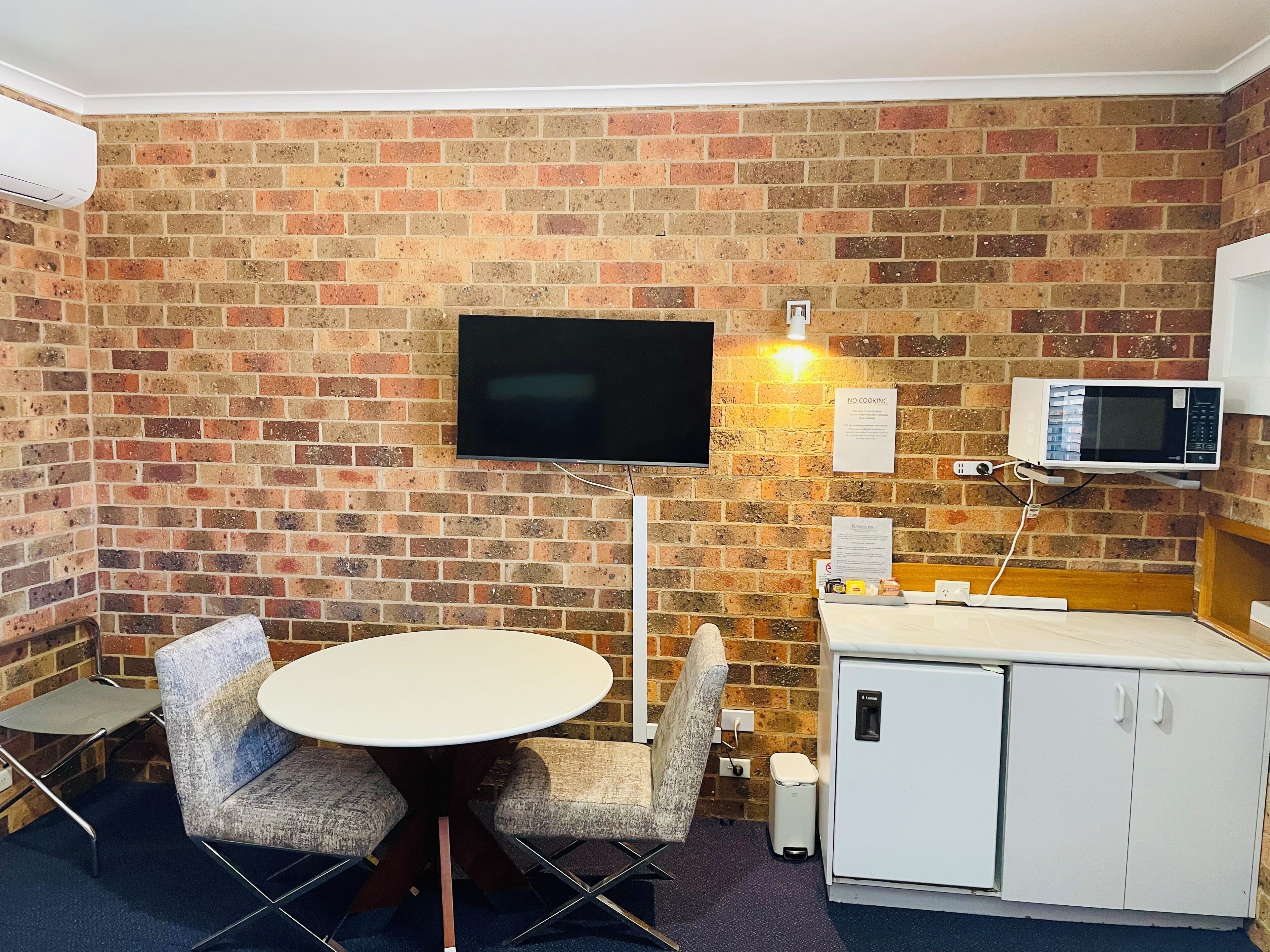 A room with a brick wall, a flat-screen TV, a microwave, a small refrigerator, and a round table with four chairs.